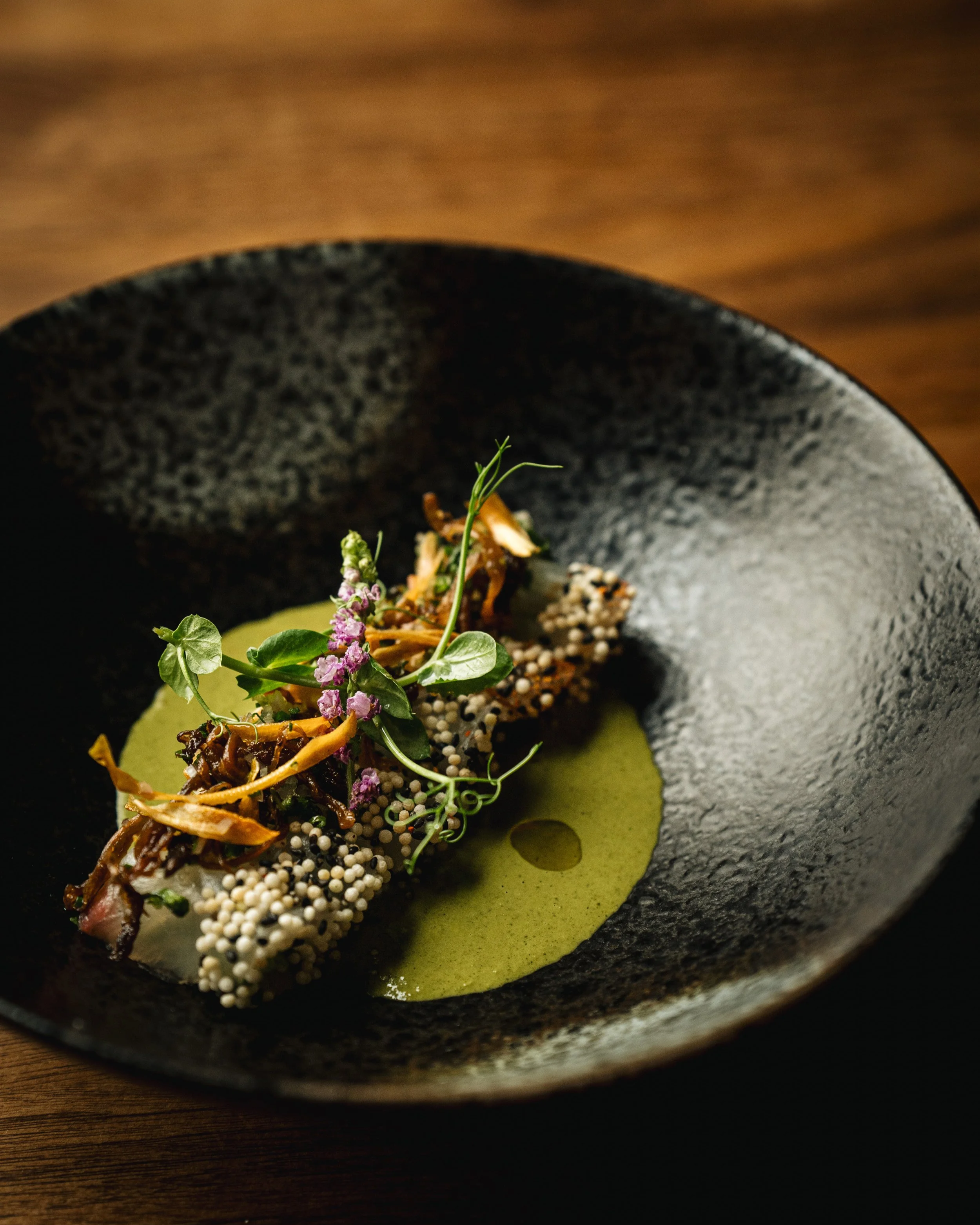 A plated gourmet dish with a green sauce, garnished with herbs, microgreens, purple flowers, and crispy toppings, served in a black textured bowl on a wooden table.