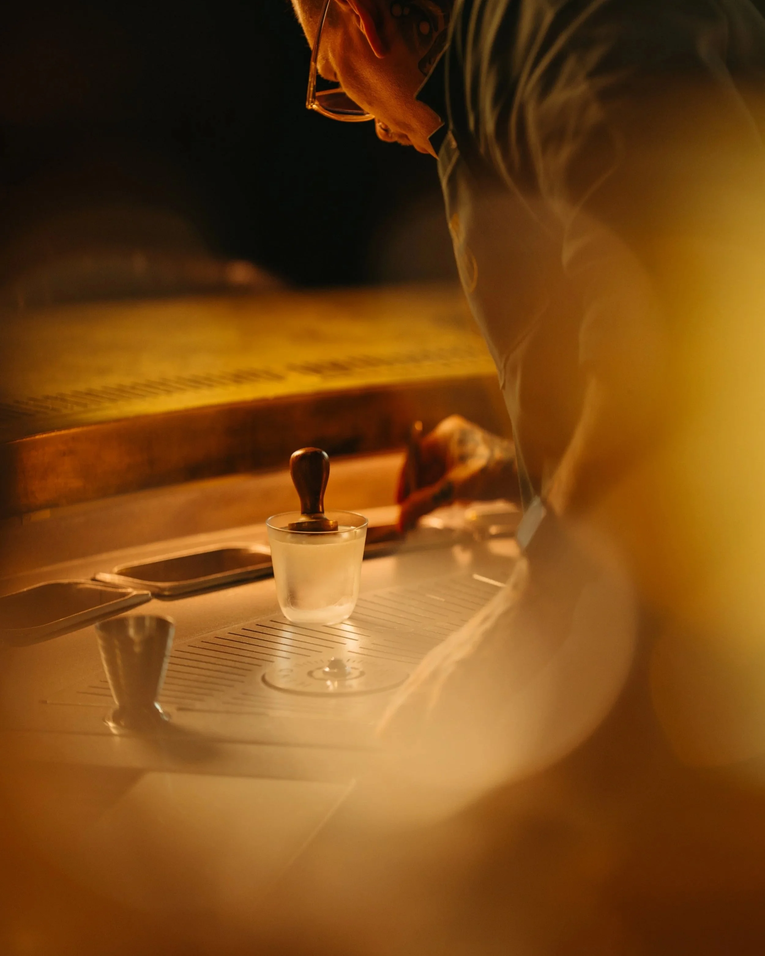 A person with glasses and tattoos on their neck, wearing a striped shirt, is leaning over a table with a candle and some equipment, in a warm, dimly lit environment.