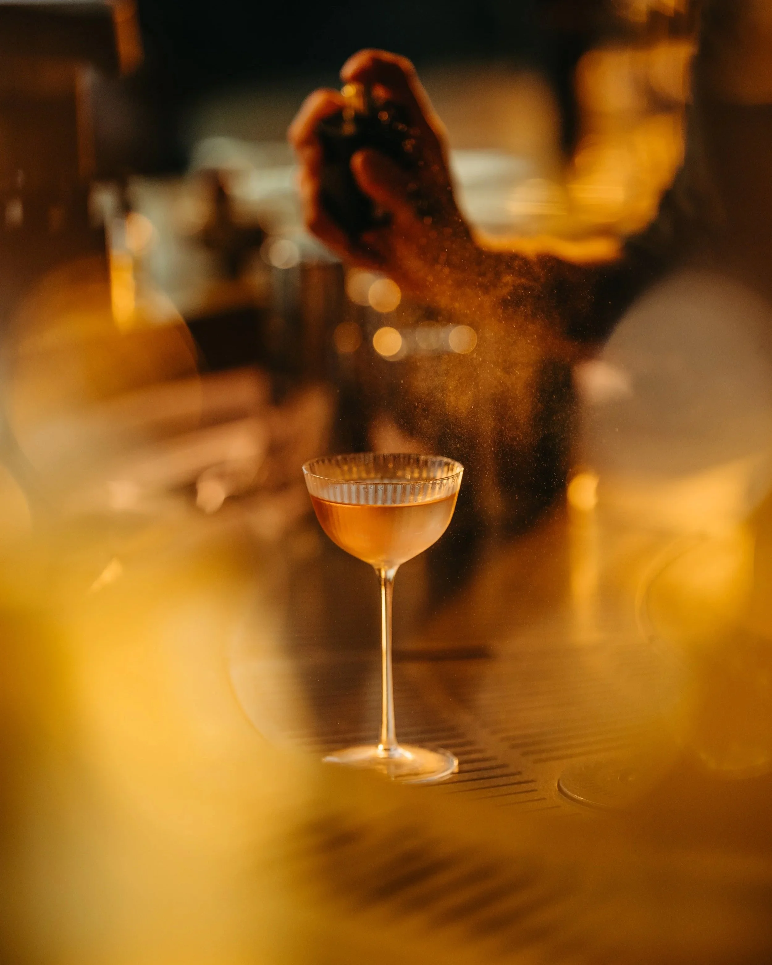 A bartender is shaking a cocktail shaker behind a bar counter at night, with a martini glass in the foreground.