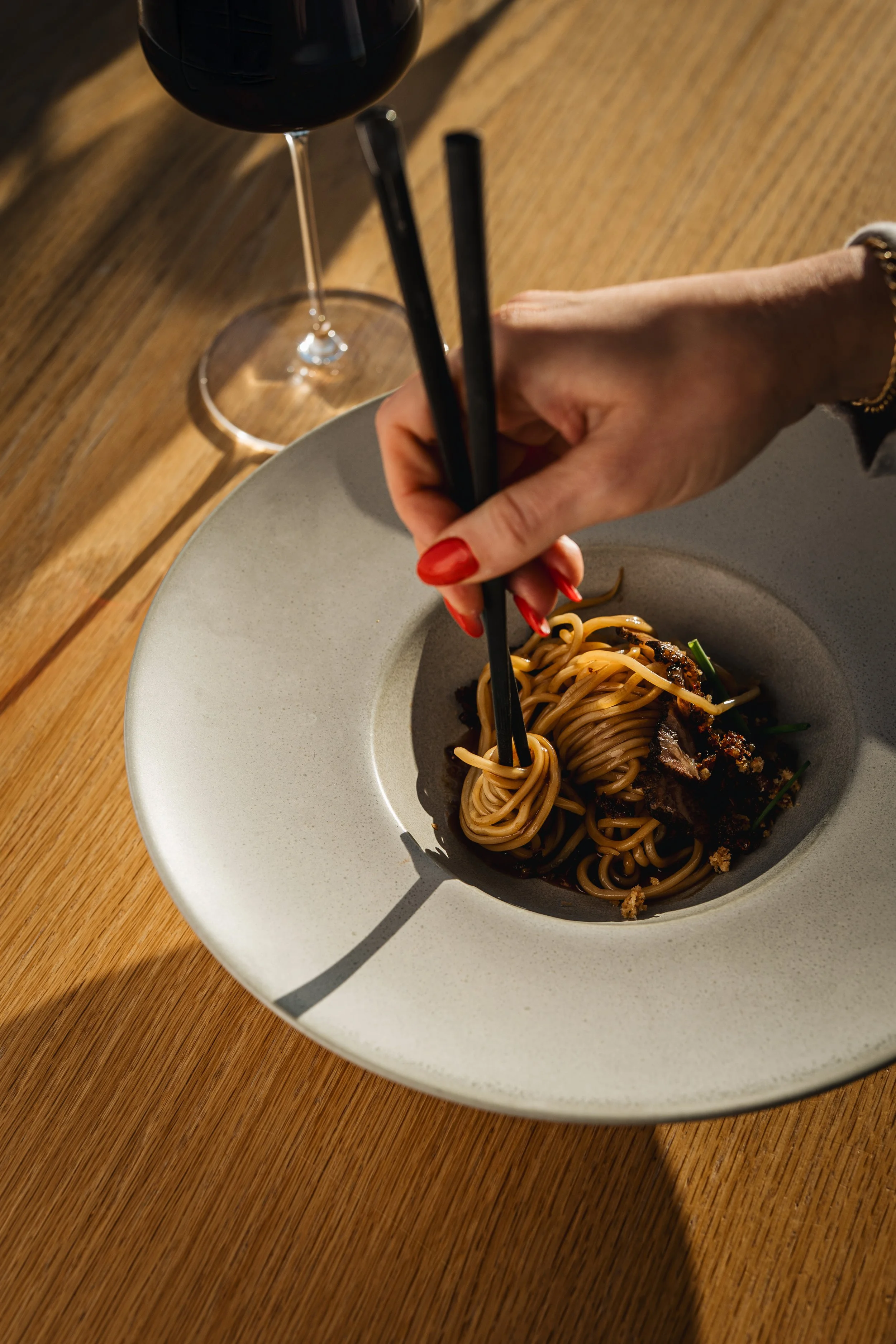 A person using chopsticks to pick up noodles from a bowl of Asian cuisine on a wooden table, with a glass of red wine nearby.