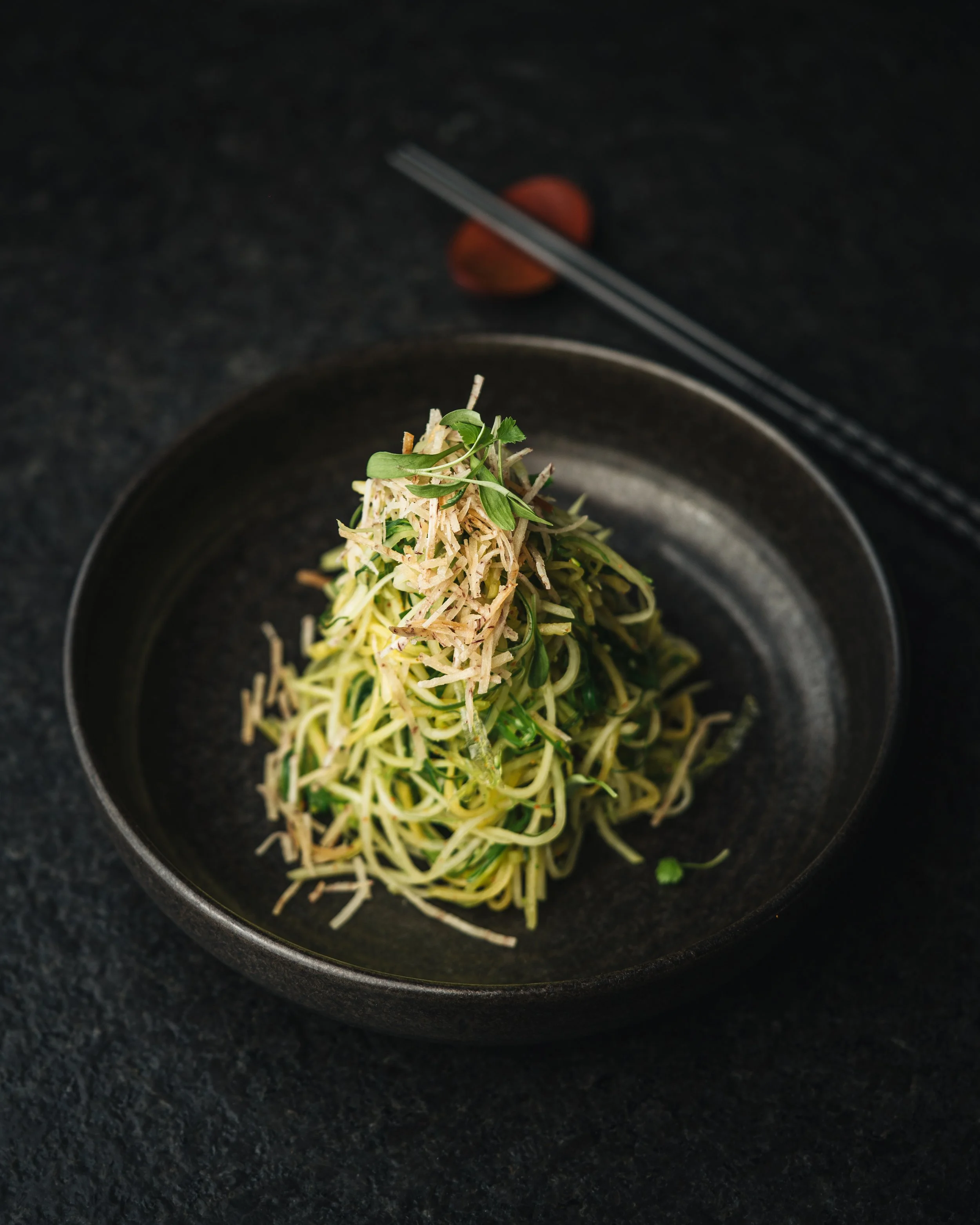 Close-up of a black bowl with spiralized zucchini noodles garnished with microgreens, on a black countertop.