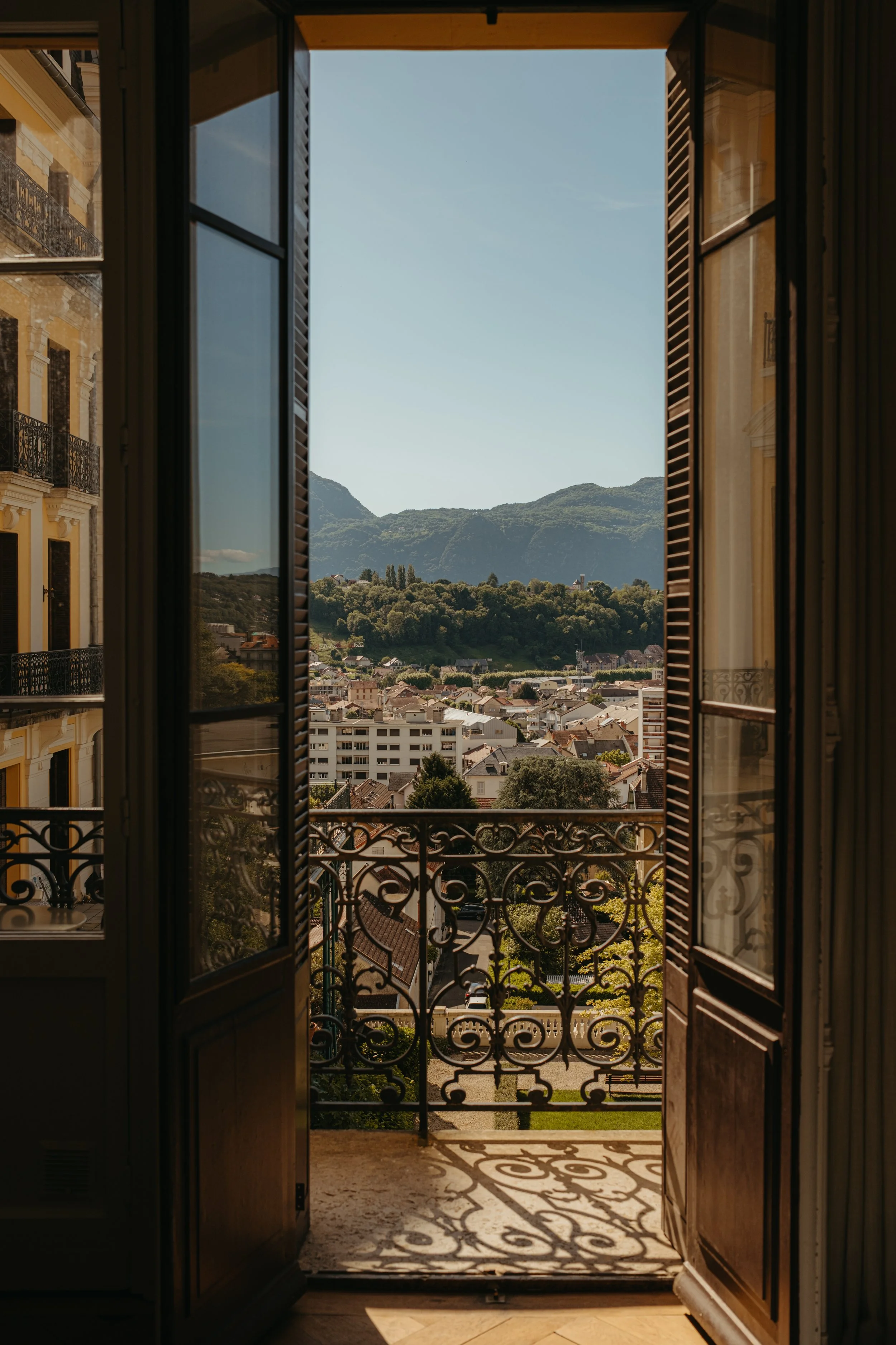 View from an open window showing city rooftops, lush green trees, a hillside with houses, and mountains in the background under a clear blue sky.