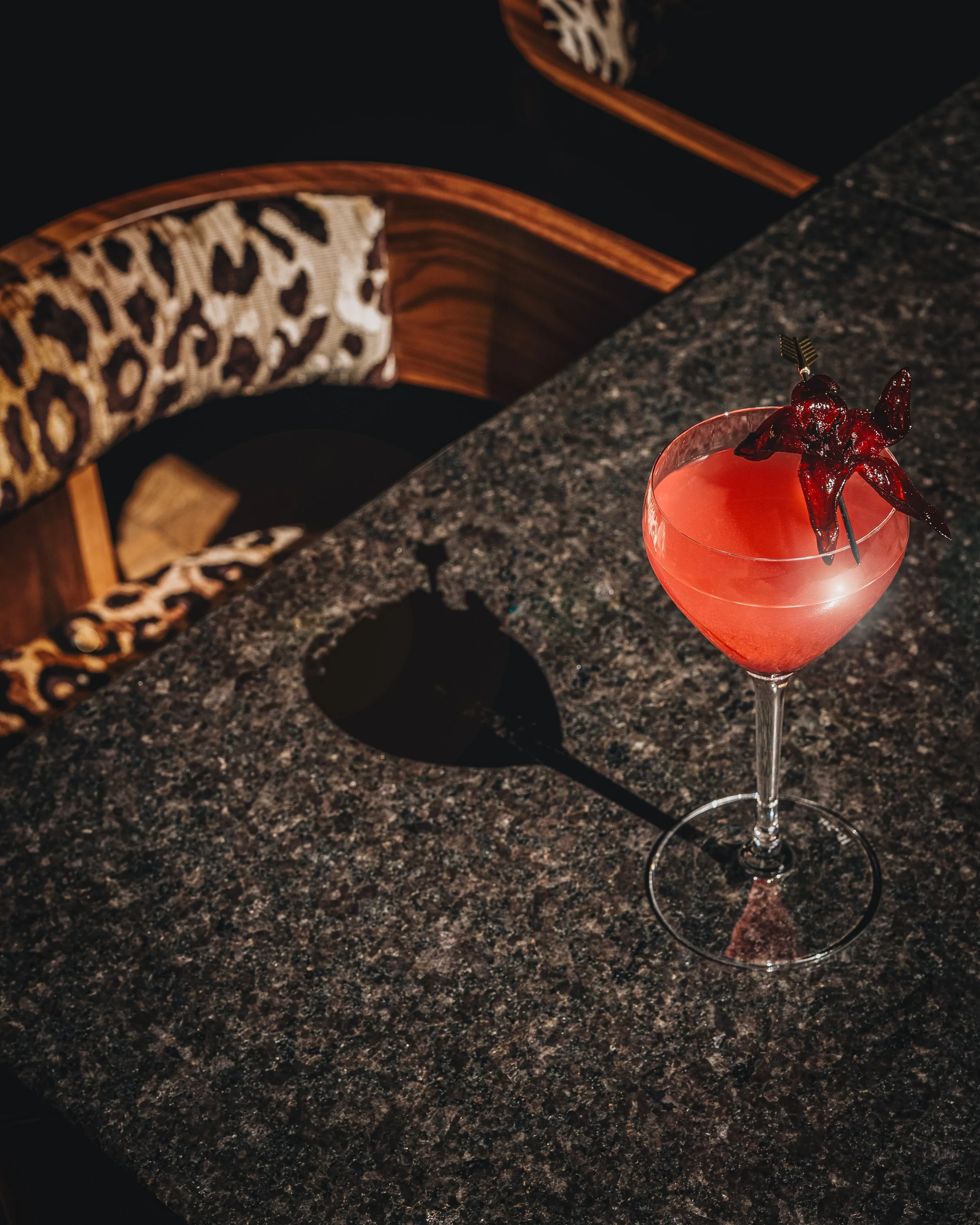 A pink cocktail in a coupe glass garnished with a dark red cherry, a star-shaped fruit, and a tiny fork, sitting on a dark granite countertop with a leopard-print upholstered chair and a wooden backrest visible in the background.