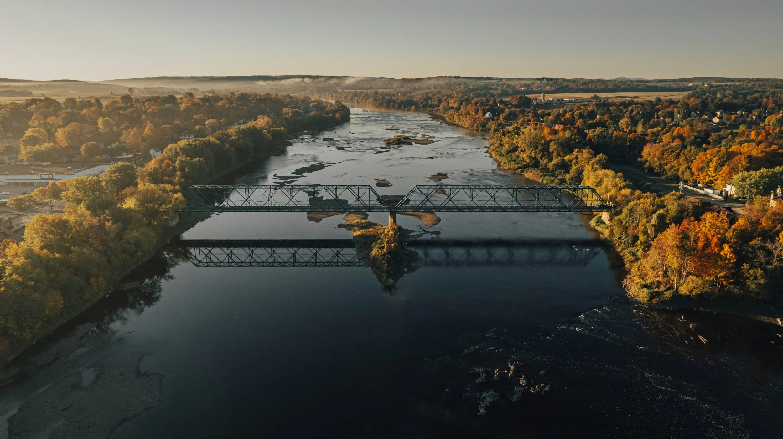 An aerial view of a river with bridges during fall, surrounded by trees with colorful autumn foliage and a small town in the distance.