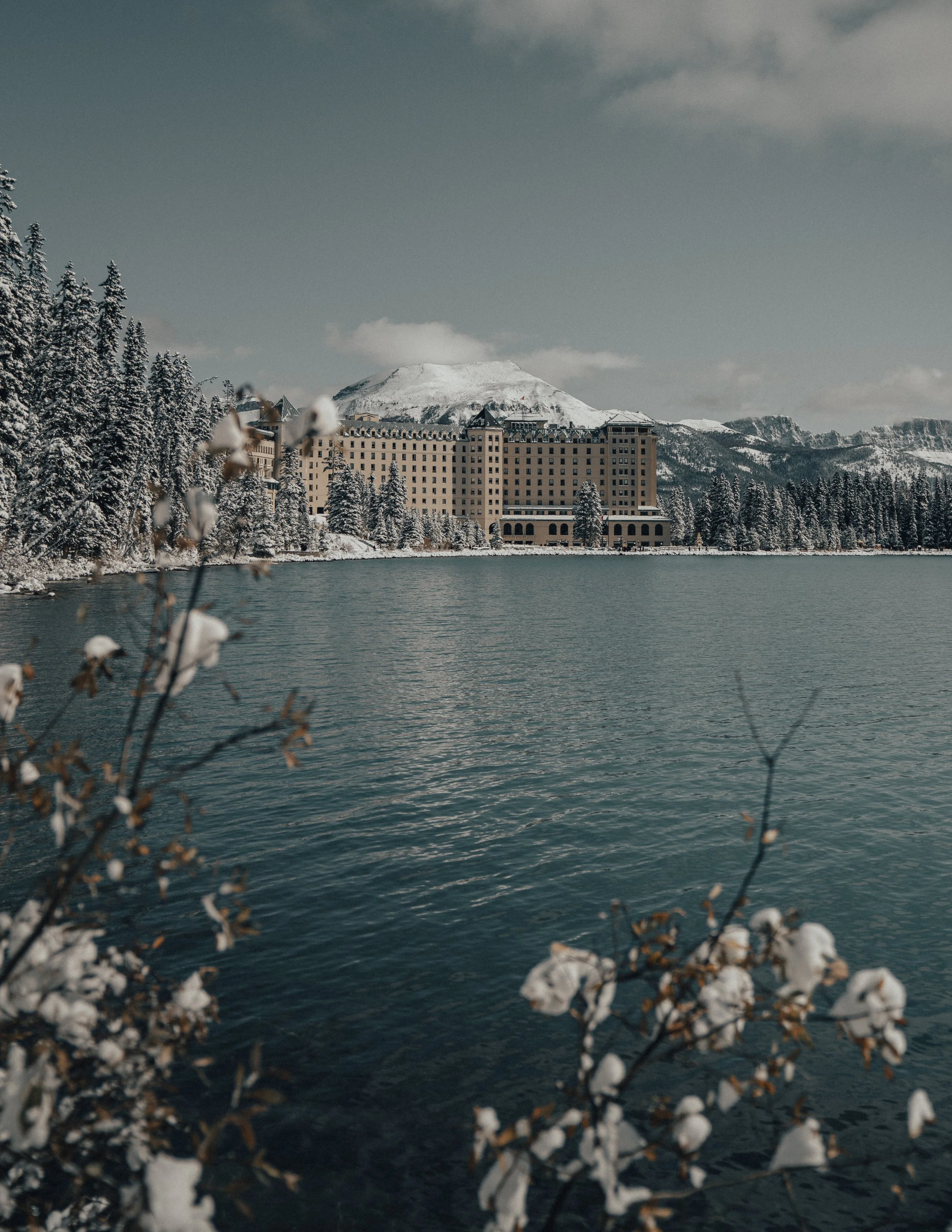 A snow-covered mountain with a large hotel or resort building at its base, trees covered in snow along the water's edge, and a lake or body of water in the foreground.