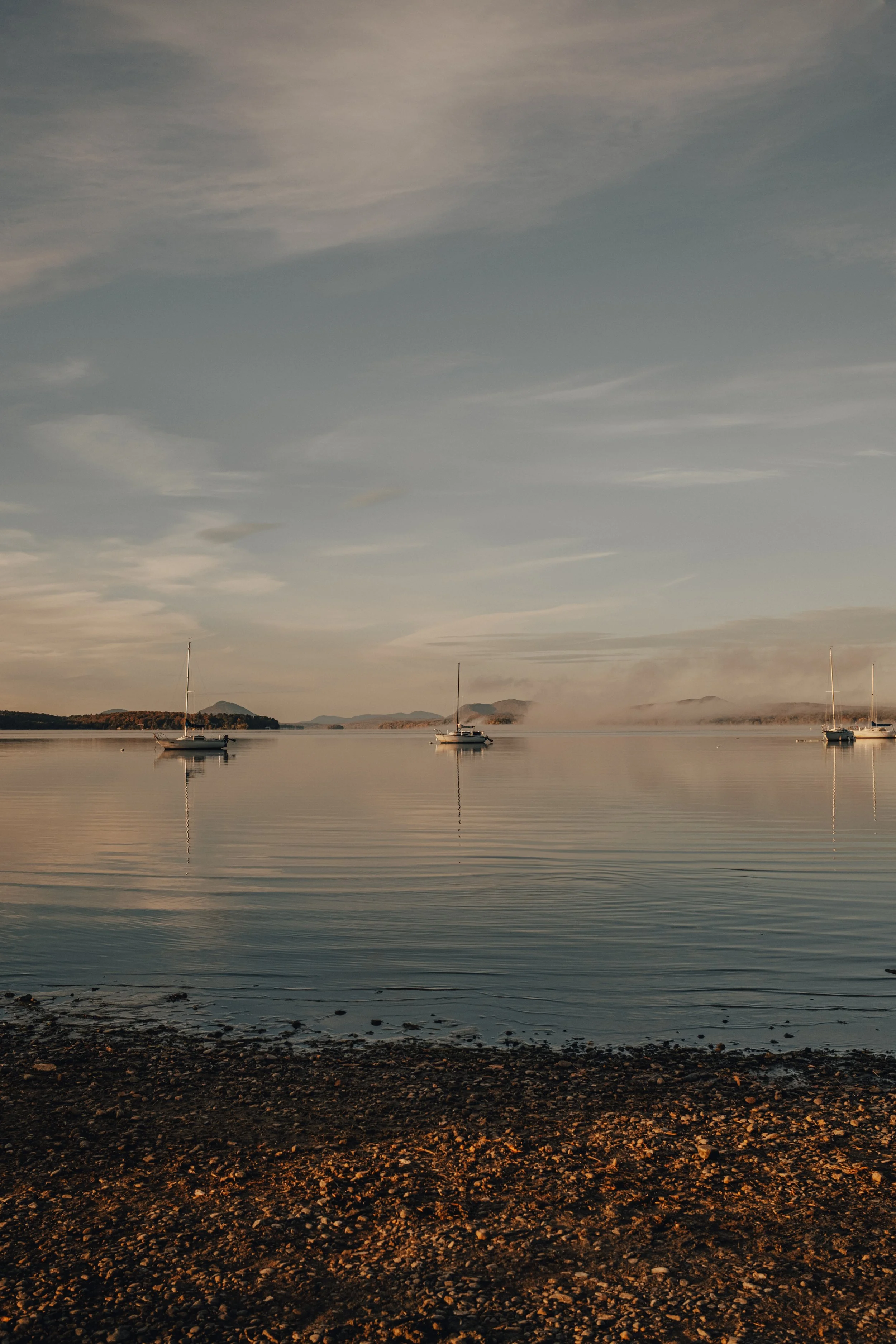 Three sailboats anchored in calm water near a rocky shoreline under a cloudy sky.