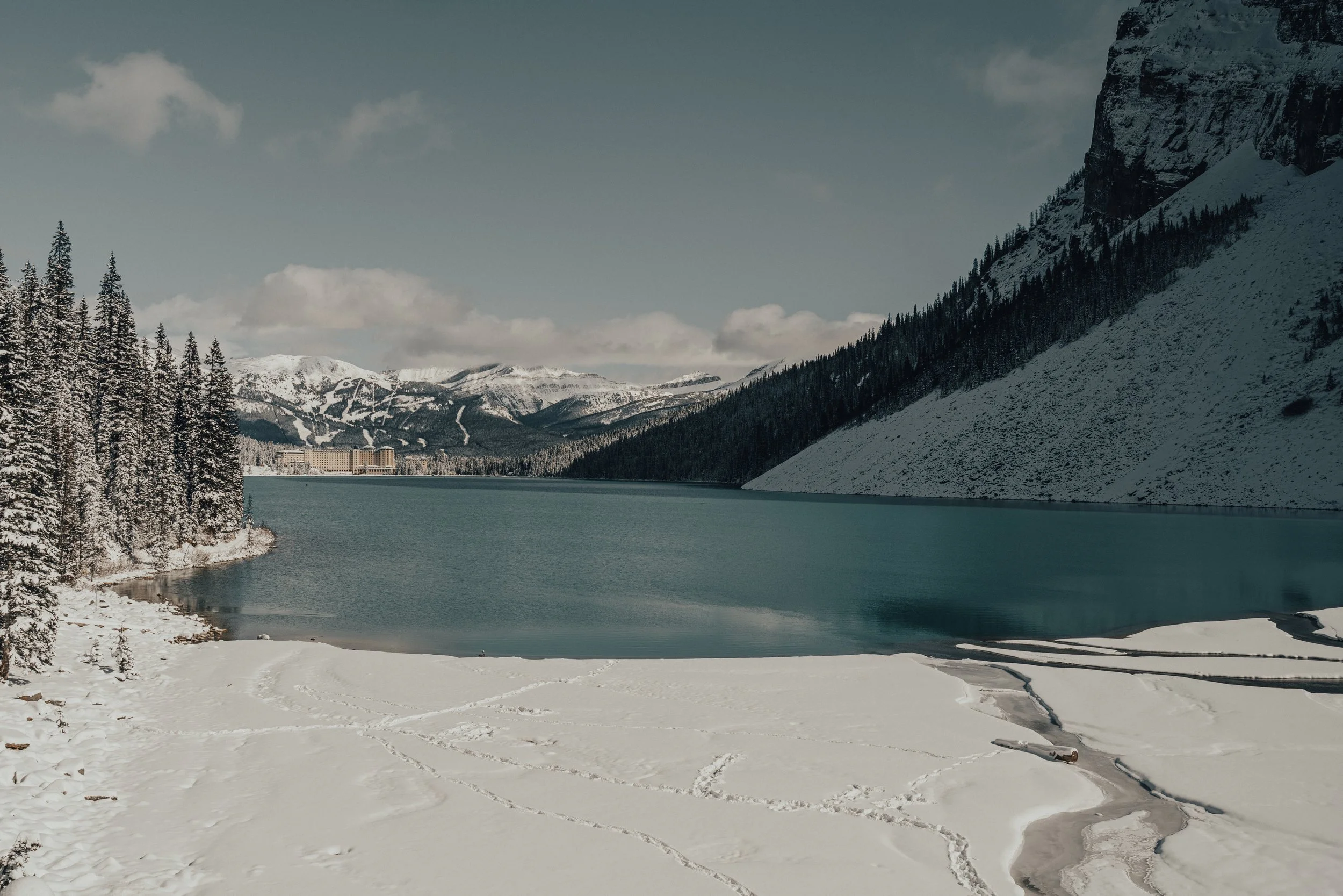 Snow-covered shoreline with a lake, mountains in the background, and a hotel or resort building near the water, with skis and ski tracks on the snow.