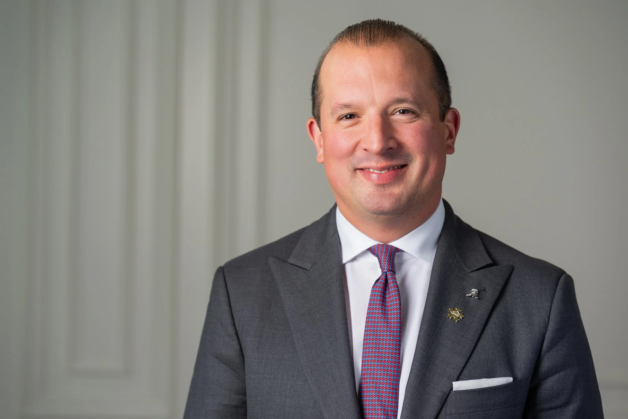 A man in a dark gray suit with a white shirt and a colorful tie, smiling in front of a plain background.