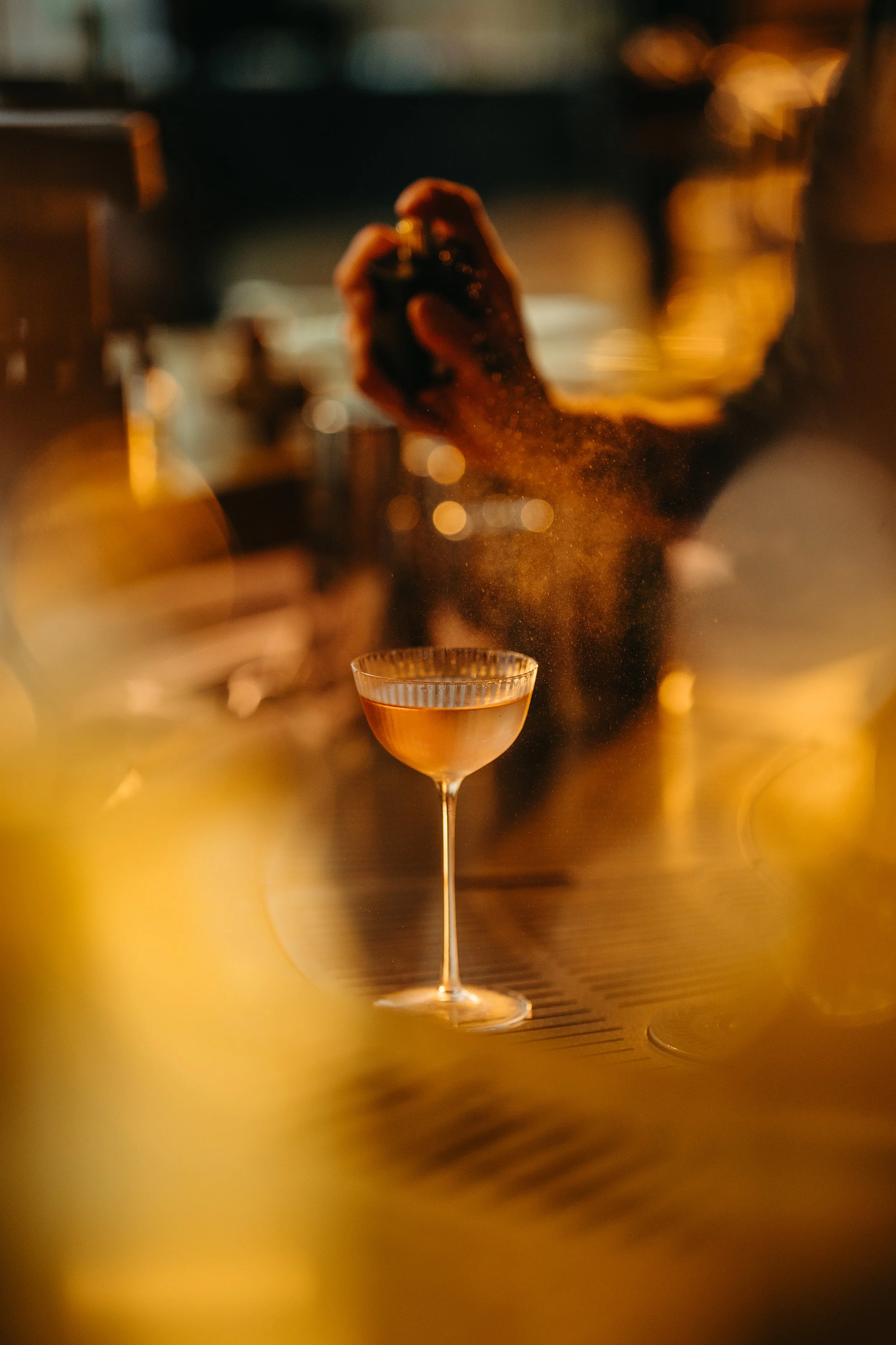 A bartender is shaking a cocktail shaker behind a bar counter at night, with a martini glass in the foreground.