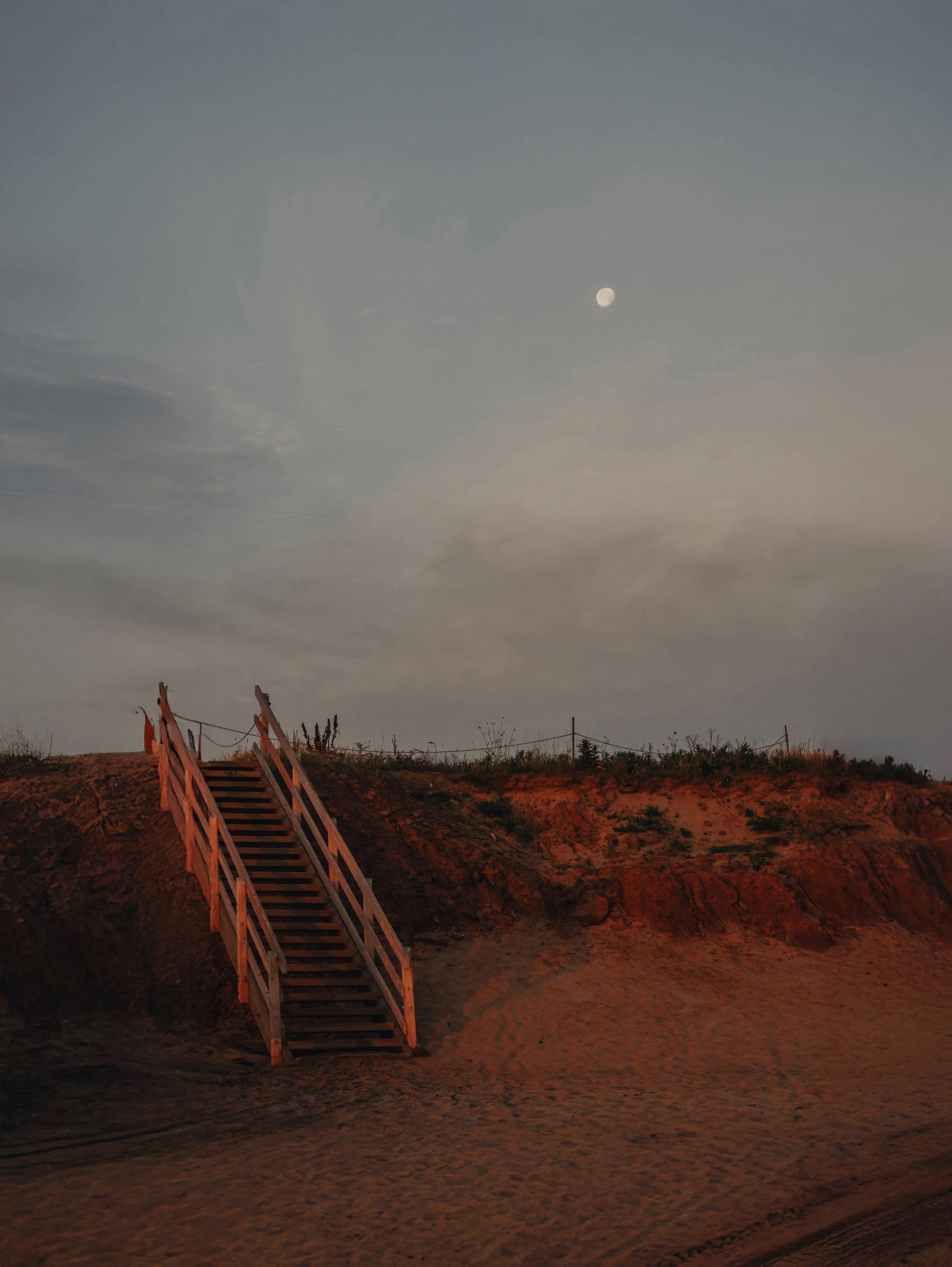 Early evening scene of a dirt pathway with a wooden staircase leading up to a hill. The sky is partly cloudy with a bright moon visible.