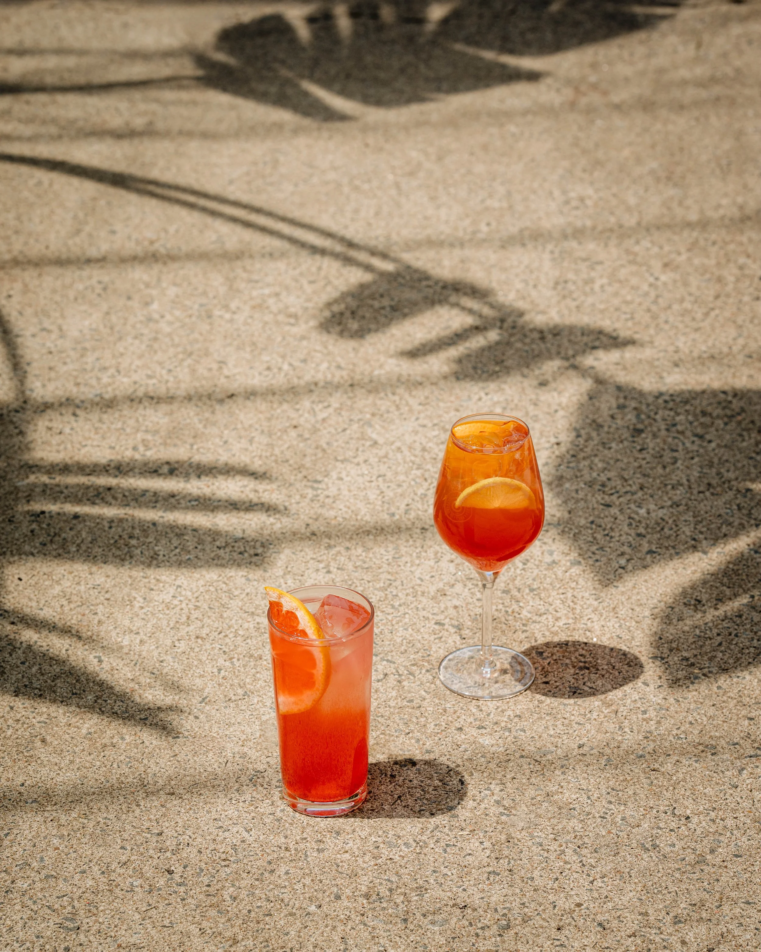 Two cocktails, a tall glass of red/orange drink with an orange slice and ice, and a shorter glass with orange beverage garnished with orange slices, sitting on a sandy surface with shadows cast across.