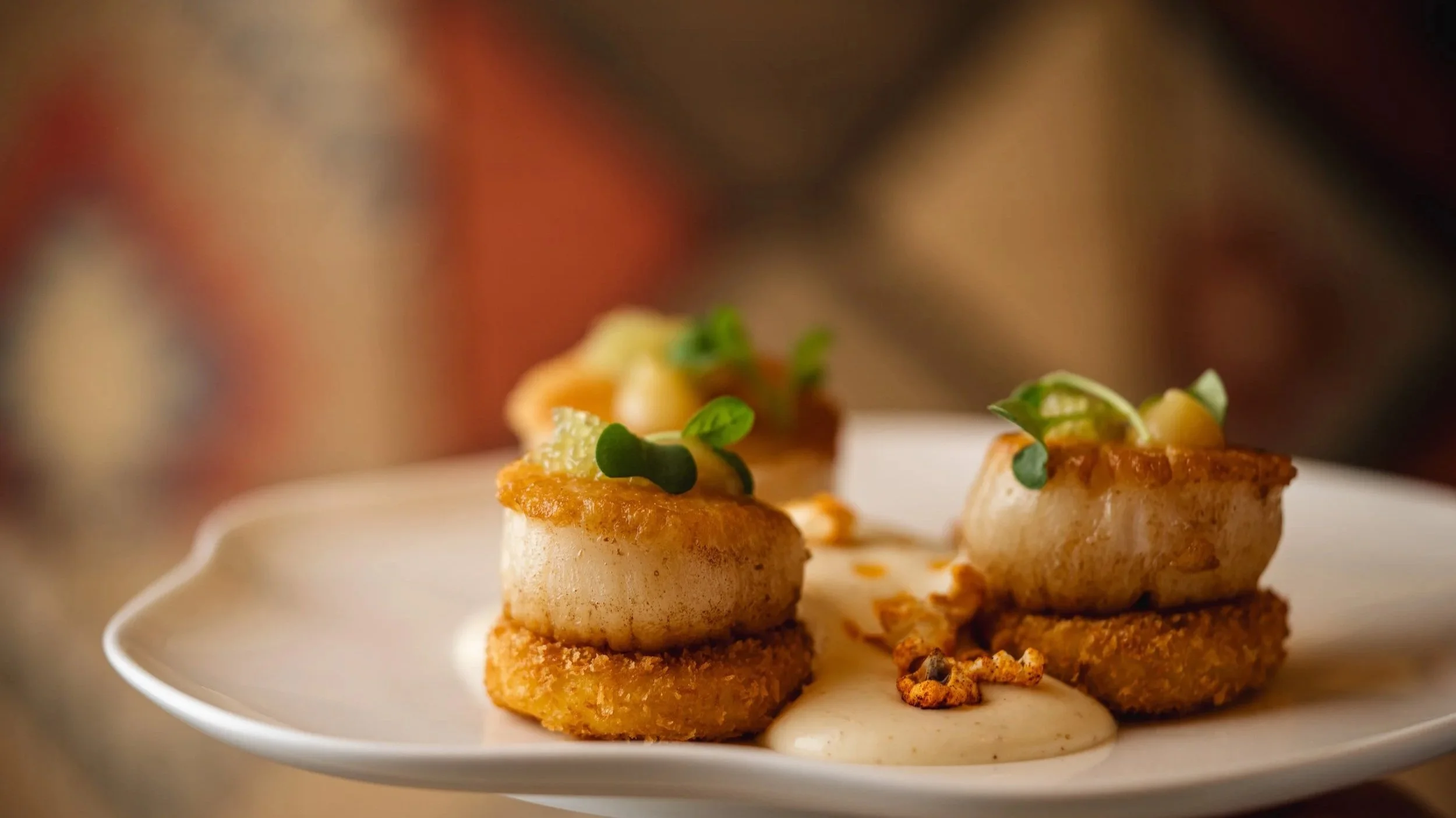 Close-up of three seared scallops topped with herbs and sauce, plated on a white dish.