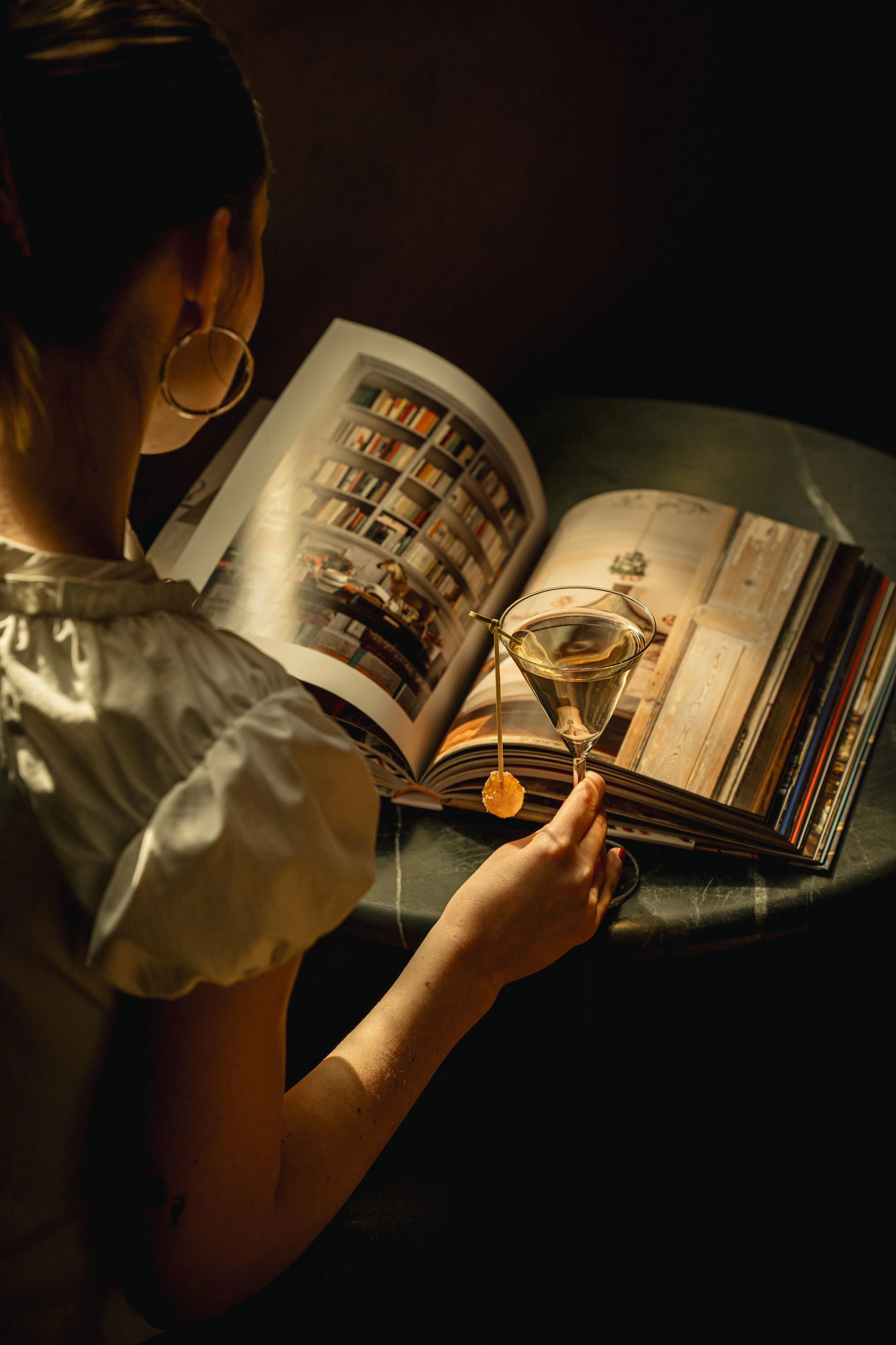 A woman with hoop earrings reading a large art book at a dark table, holding a cocktail glass with a cherry garnish in a dimly lit setting.