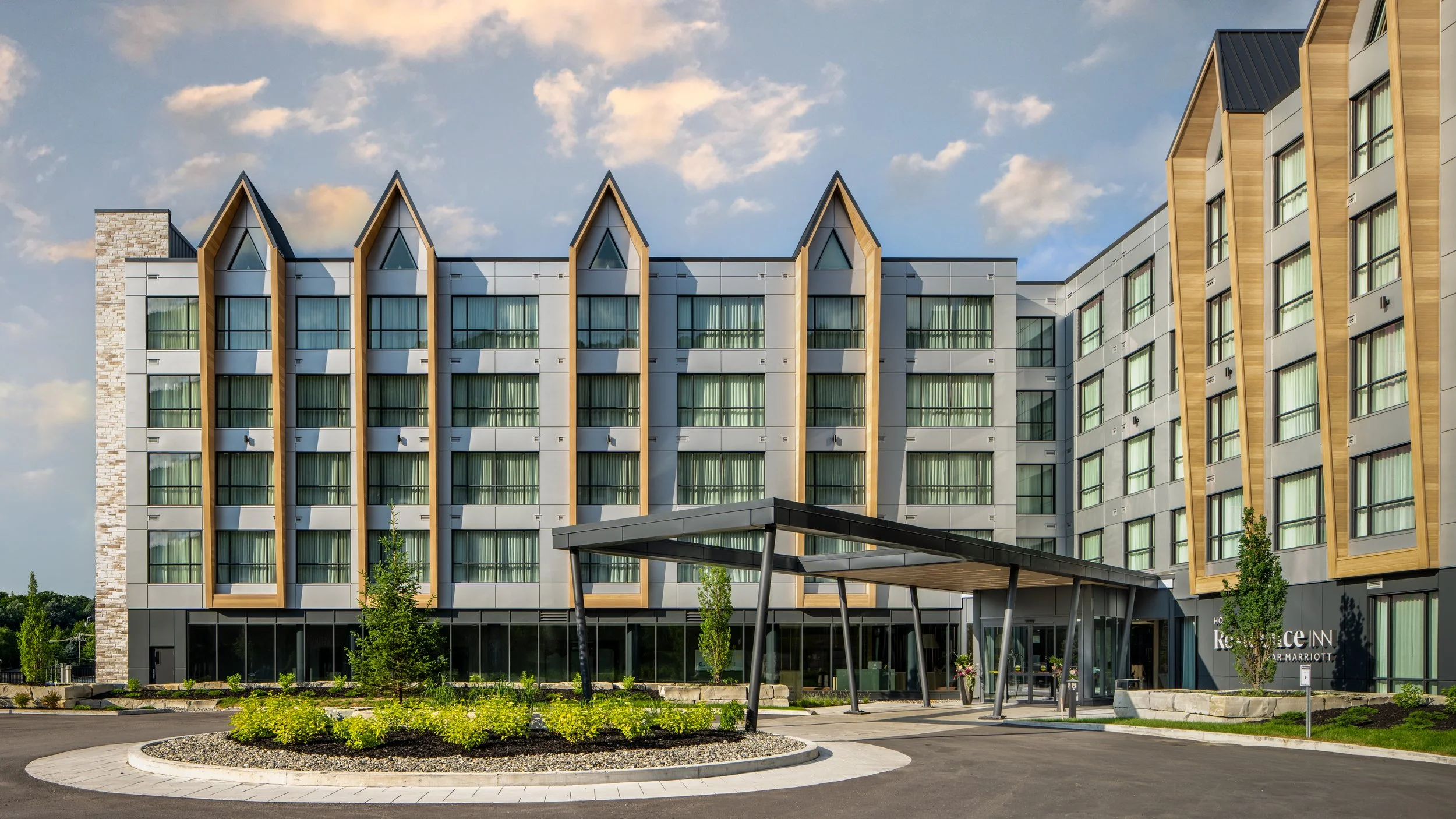 Modern hotel building with a glass and wood facade, featuring pointed roof peaks, glass windows, a covered entrance, and landscaped surroundings under a partly cloudy sky.