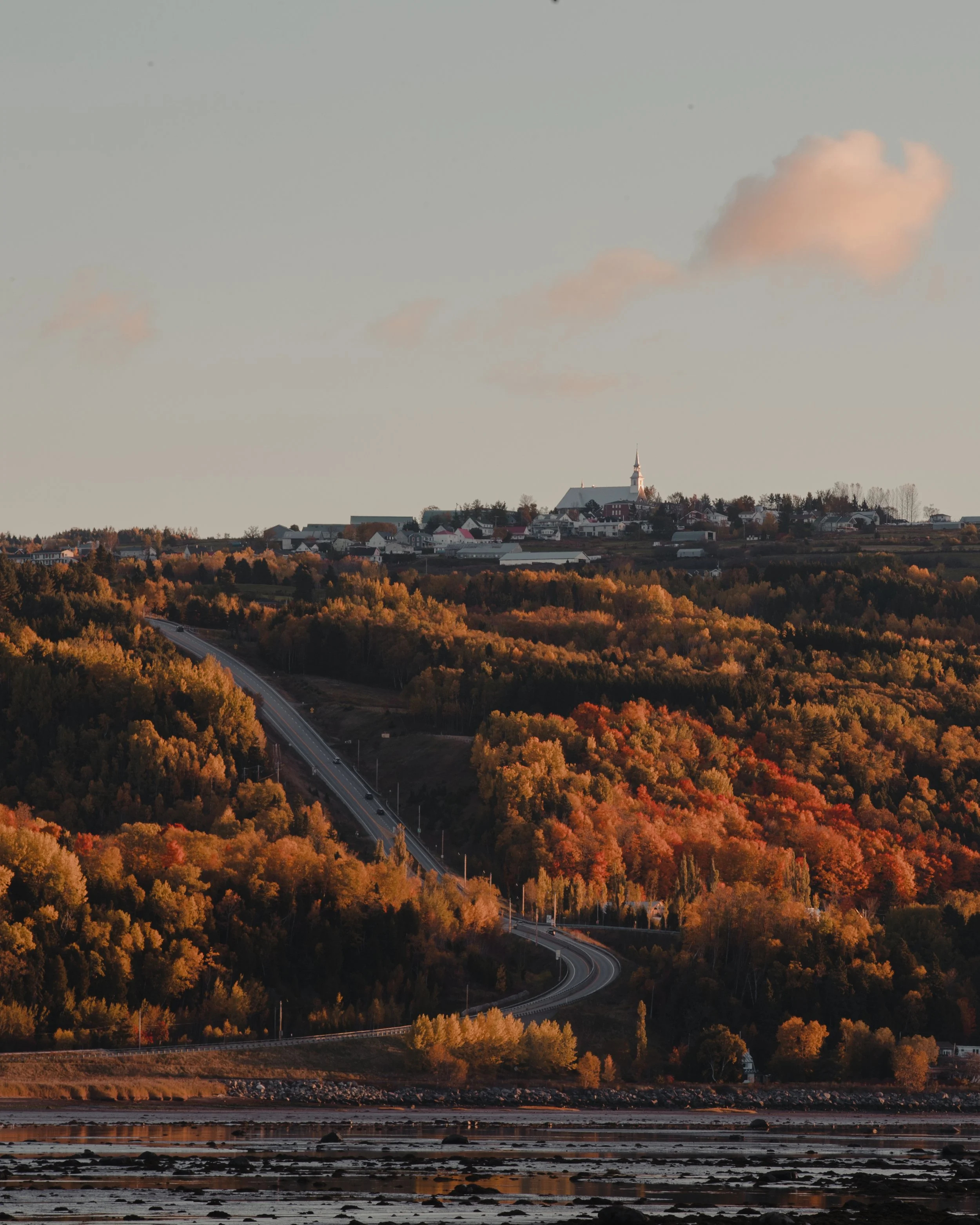 A scenic view of a small town on a hilltop with a church, surrounded by autumn-colored trees, seen at sunset with a winding road leading through the landscape and reflecting water in the foreground.