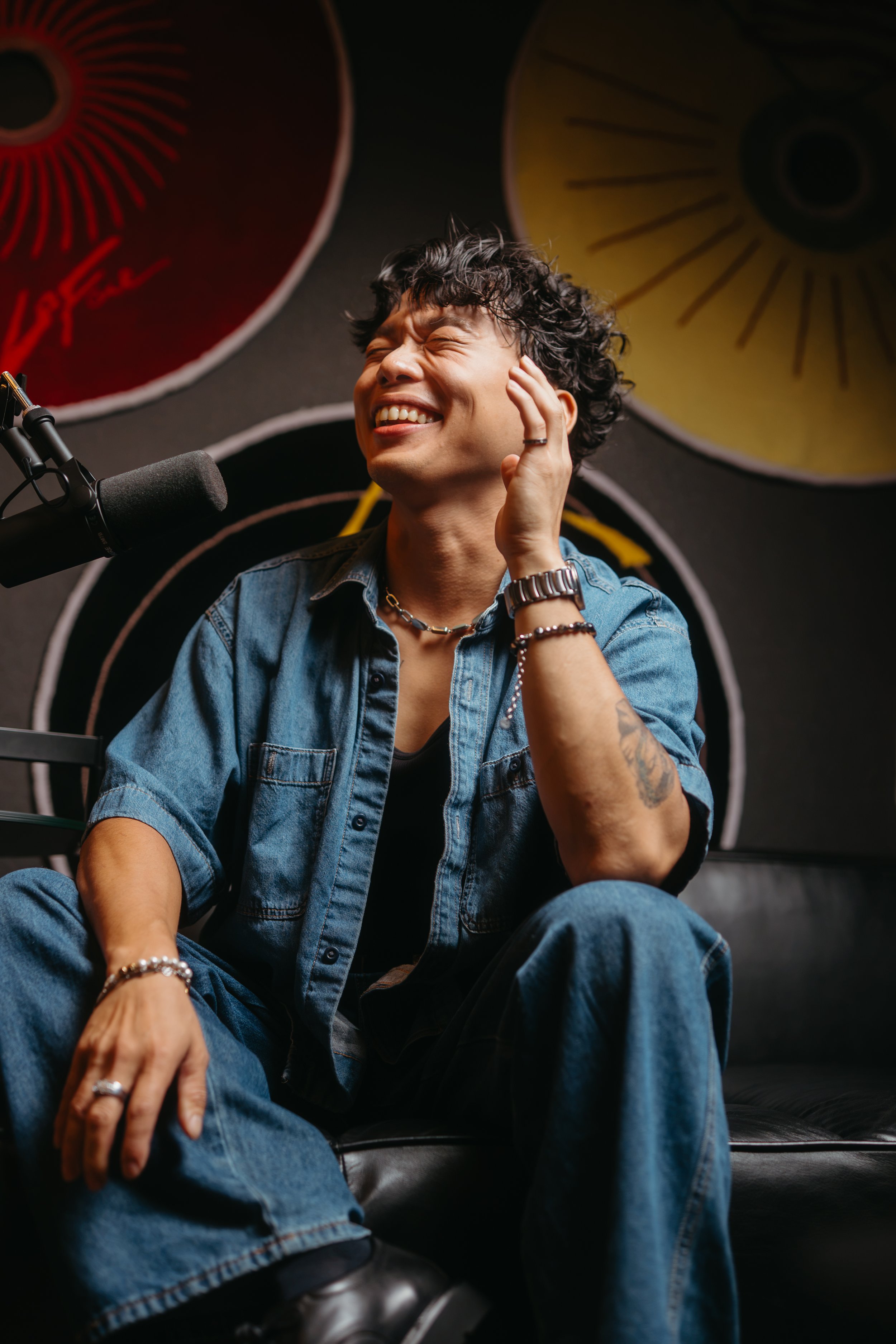 A young man with curly hair smiling and squinting, wearing a denim jacket and accessories, sitting in front of colorful circular wall art.