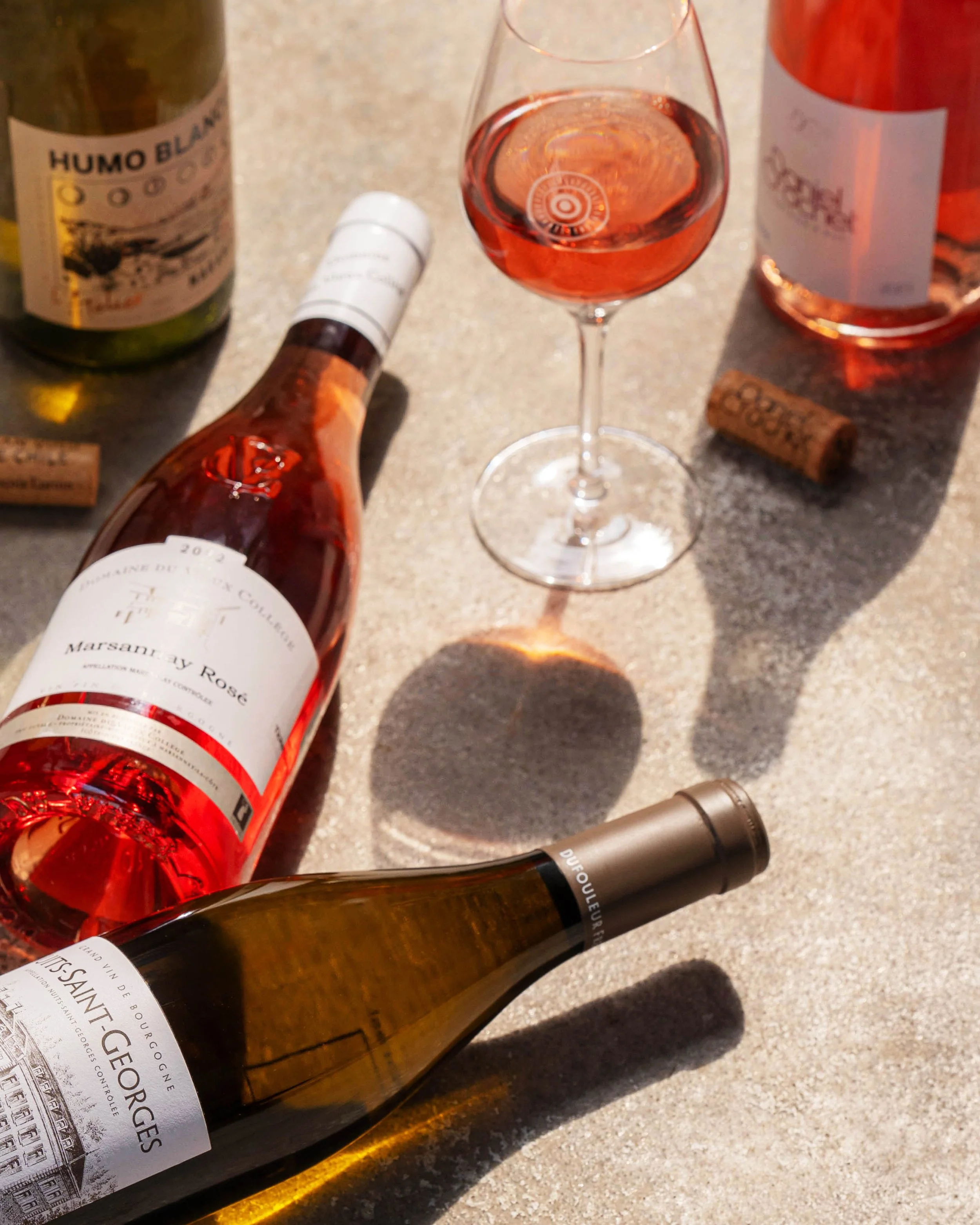 A bottle of rosé wine, a glass of rosé wine, a cork, and two other wine bottles on a concrete surface.
