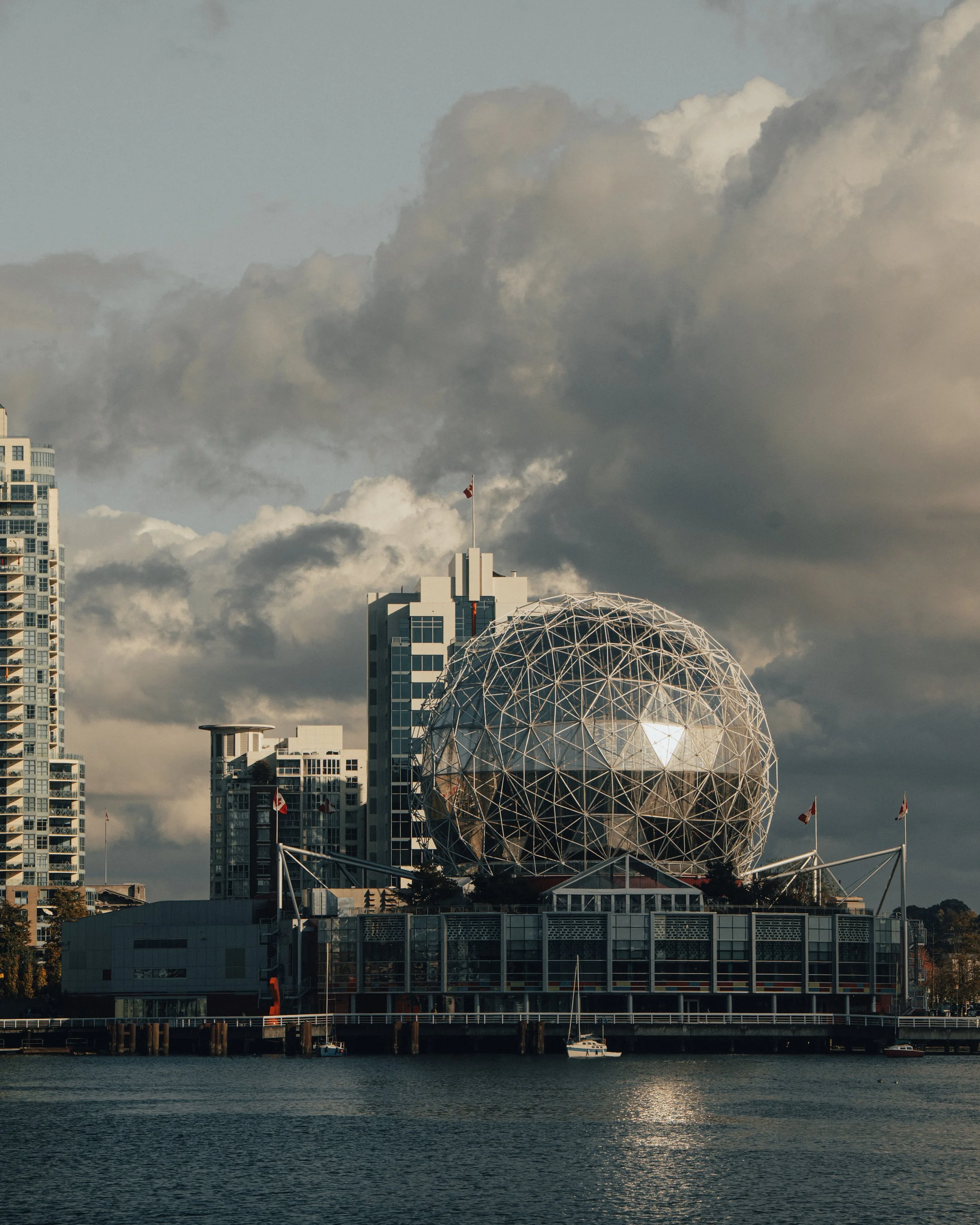 City skyline with modern buildings and a geodesic dome, with a body of water in the foreground and a cloudy sky above.