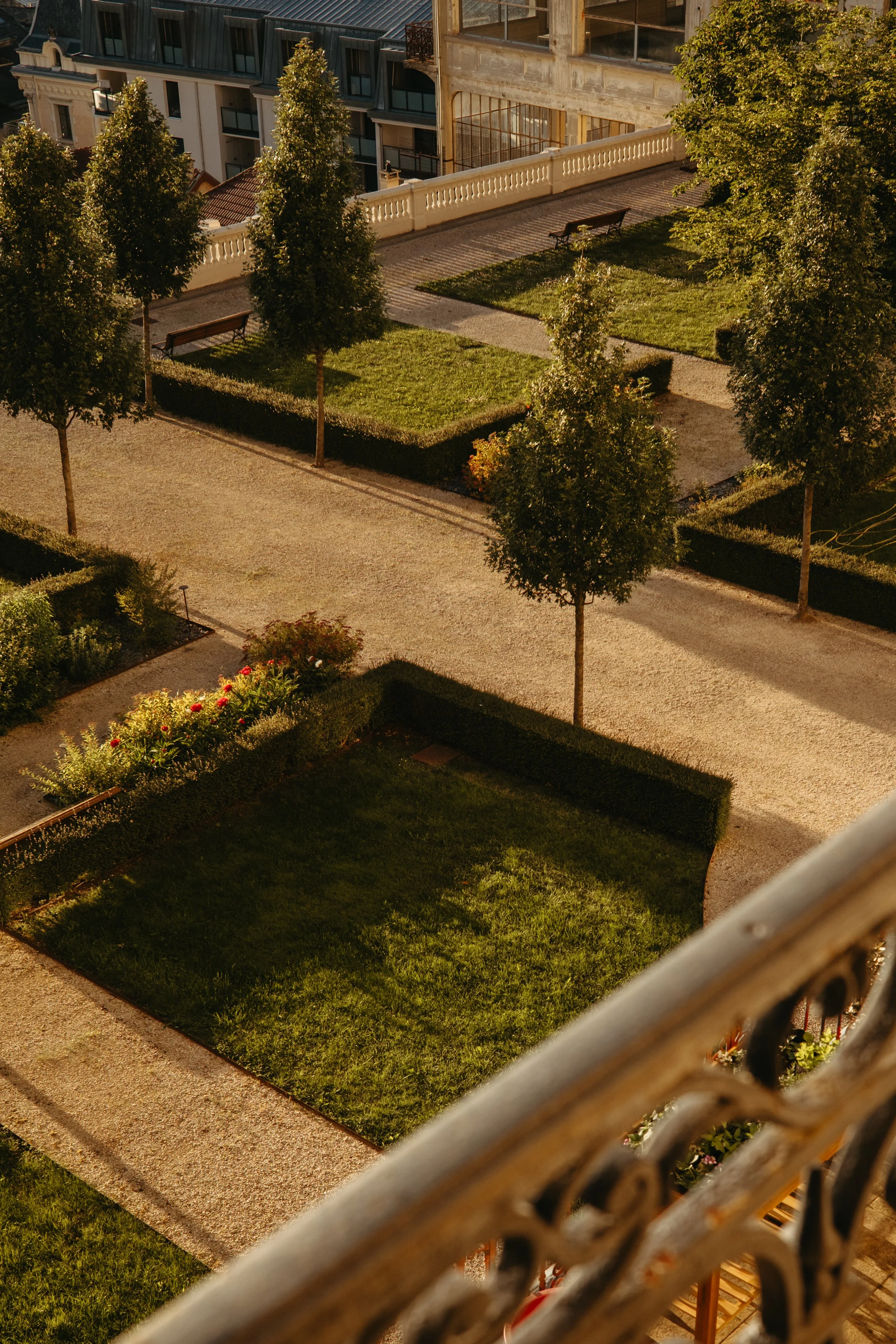 An aerial view of a small park with green grass, neatly trimmed bushes, and several trees with dense foliage. There are stone pathways, benches, and a flower bed with red flowers. The park is surrounded by buildings, some with modern architecture and