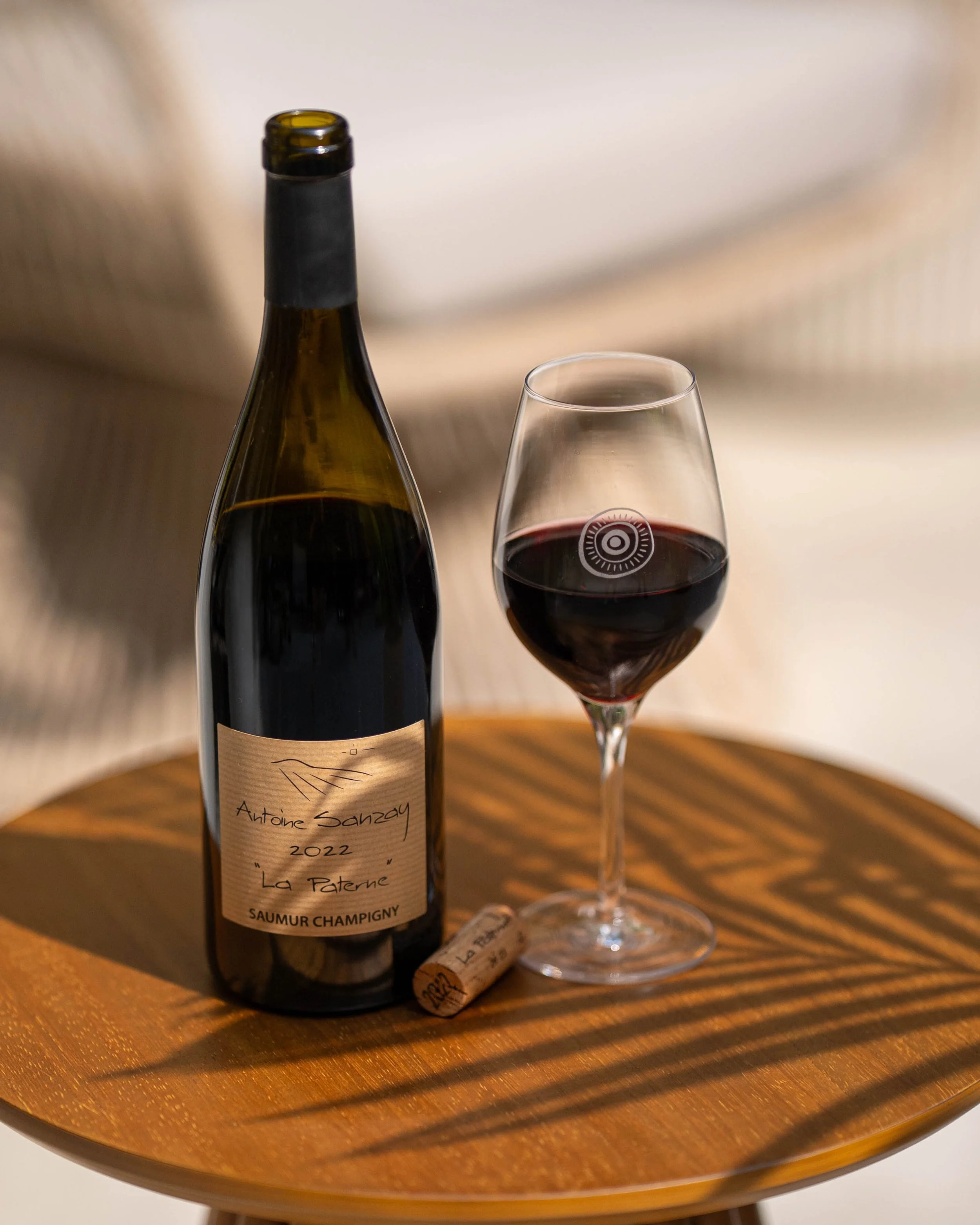 A bottle of Antoine Sanzay wine, a glass of red wine, and a cork on a round wooden table with a beige background.