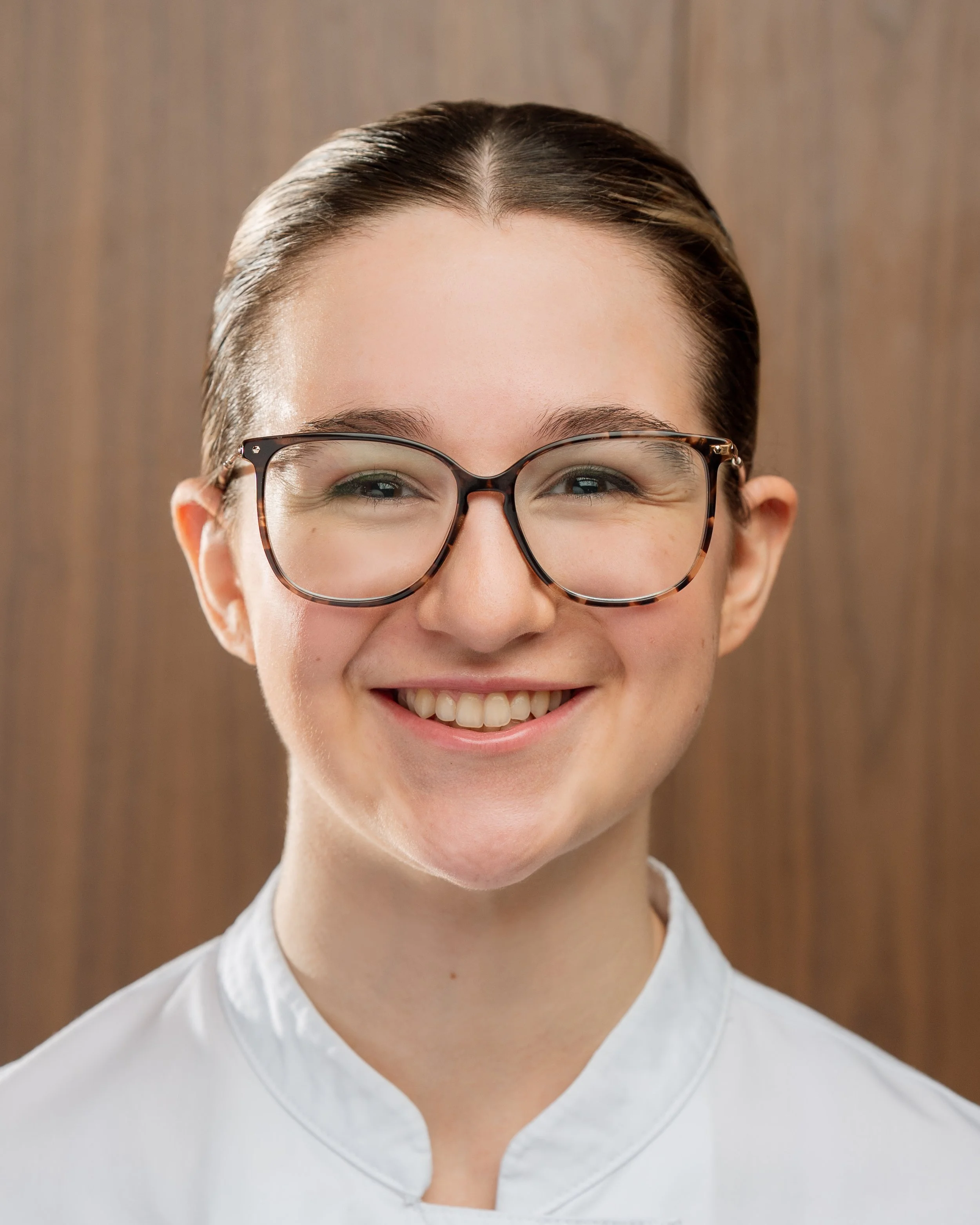 Close-up of a smiling woman wearing glasses, with hair pulled back and wearing a white shirt, against a wooden background.