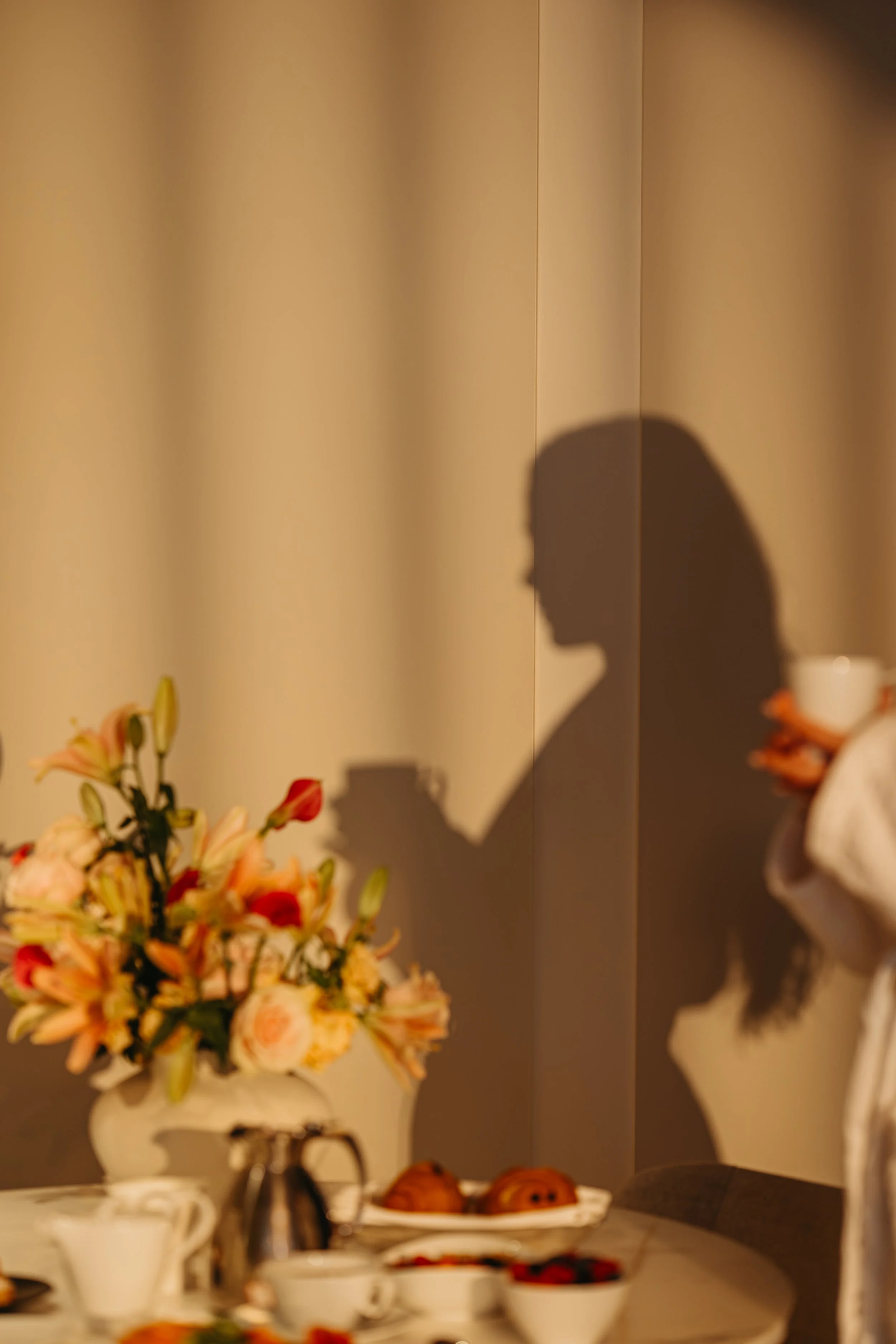 A shadow of a person on a beige wall, with a blurred floral bouquet and a table with teacups and pastries in the foreground.