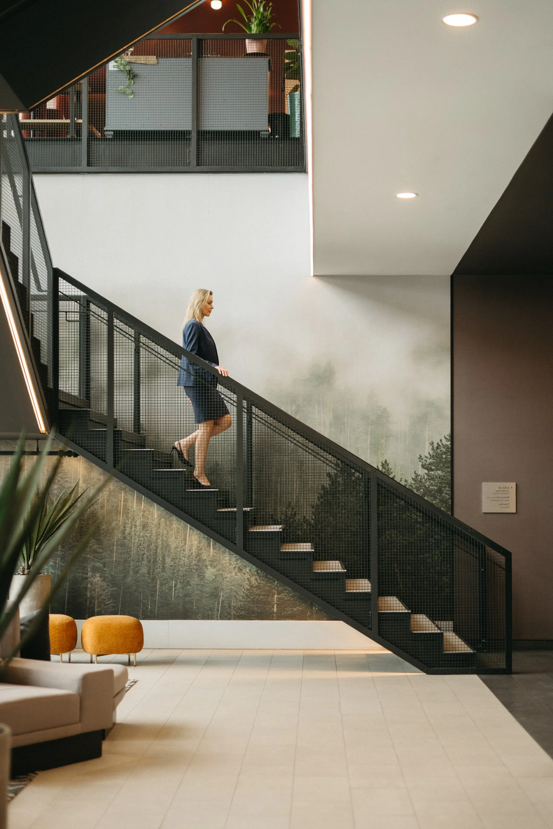 A woman in a blue suit walking down a staircase inside a modern building with minimalistic decor, a mural of a forest scene on the wall, and orange chairs in the lounge area.