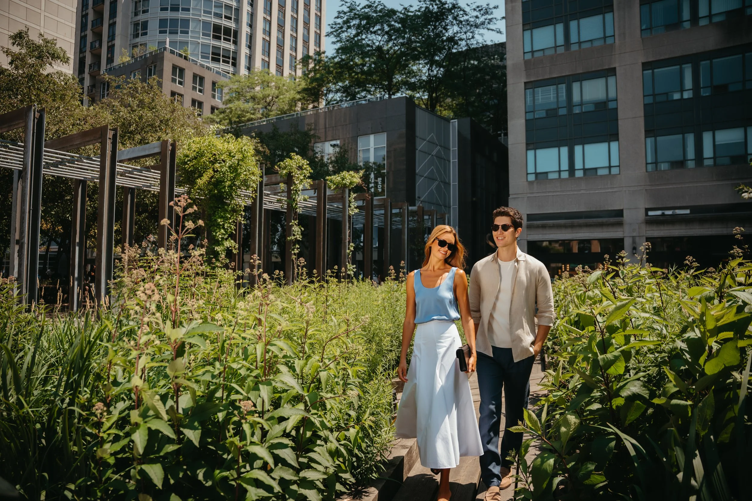A woman and a man walking together in an urban garden surrounded by tall buildings. They are smiling, wearing sunglasses, and appear to be enjoying a sunny day.
