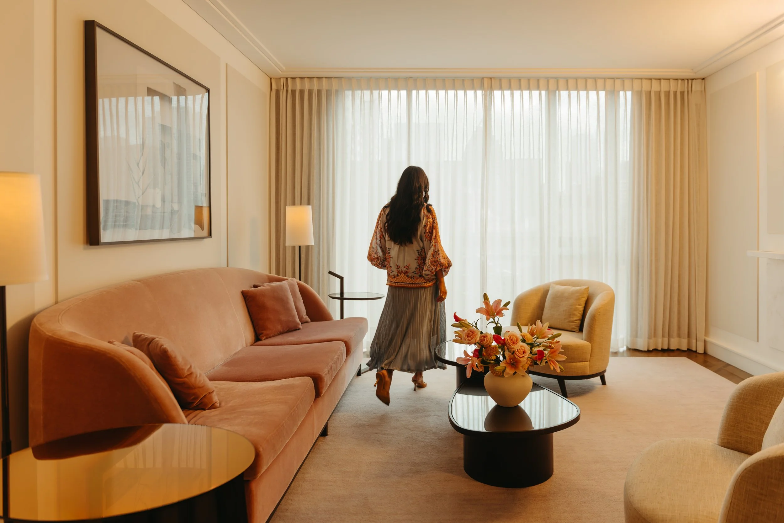 A woman with long dark hair wearing a floral top and gray skirt standing in a well-lit, modern living room with beige walls, a plush pink sofa, yellow chairs, a black coffee table with a floral arrangement, and large windows with cream curtains.