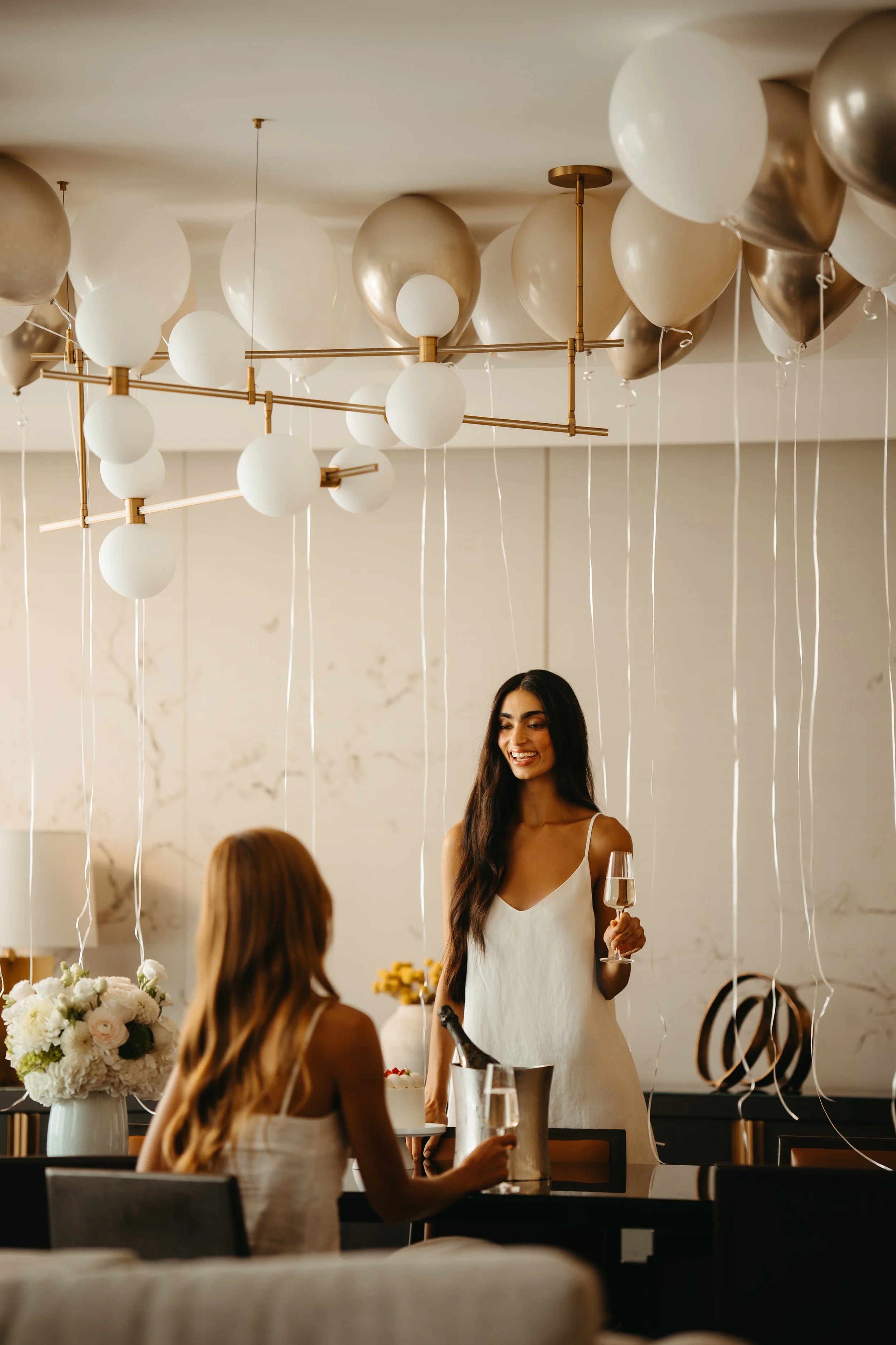 A woman in a white dress holding a glass of champagne at a celebration or party, with balloons and floral arrangements in the background.