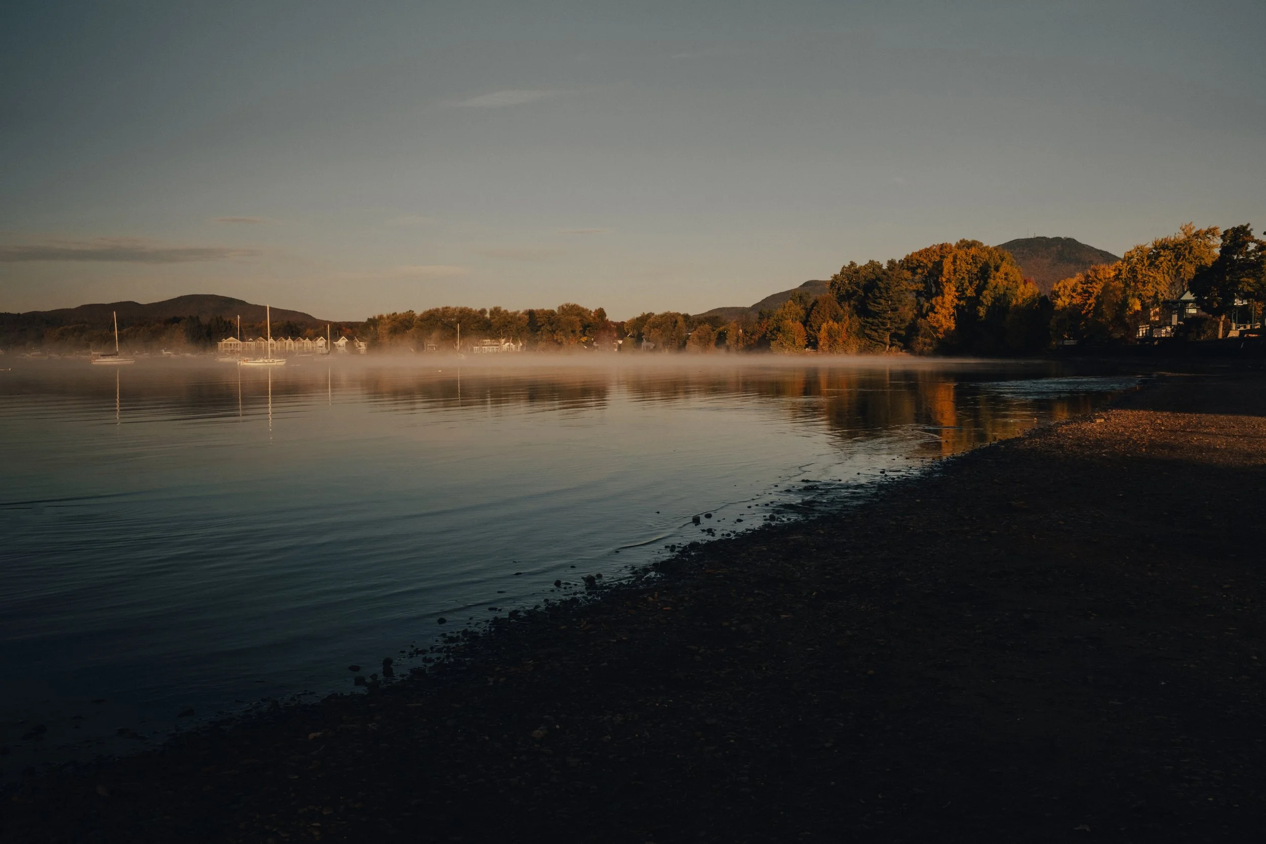 Calm lake with five sailboats docked, surrounded by mist, green trees, and distant mountains under a clear evening sky.