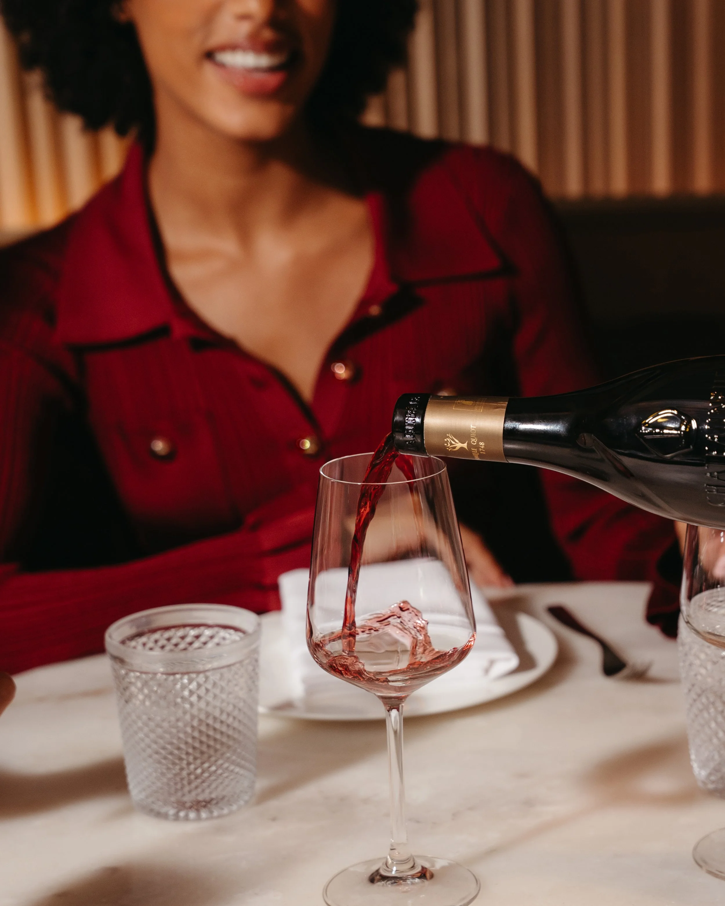 A woman in a red top pours red wine into a wine glass at a table set with glassware and plates, with a blurred background.