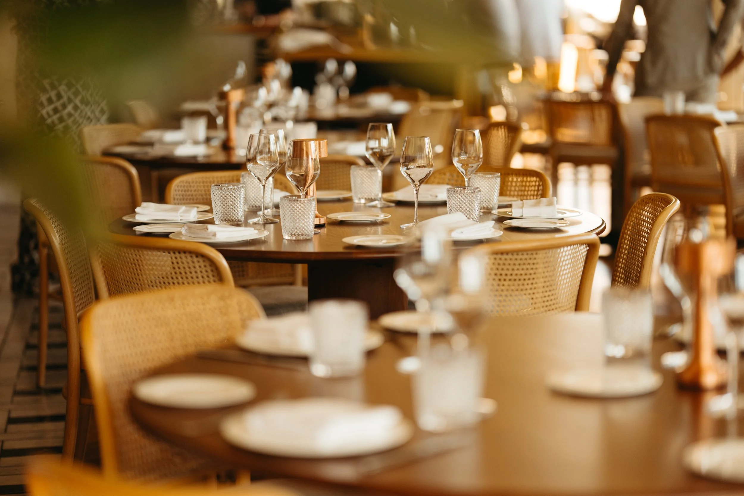 A warmly lit restaurant dining area with wooden tables set with white plates, drinking glasses, and napkins. Rattan chairs with cushioned seats surround the tables. The atmosphere appears cozy and inviting, with a blurred background of additional tab
