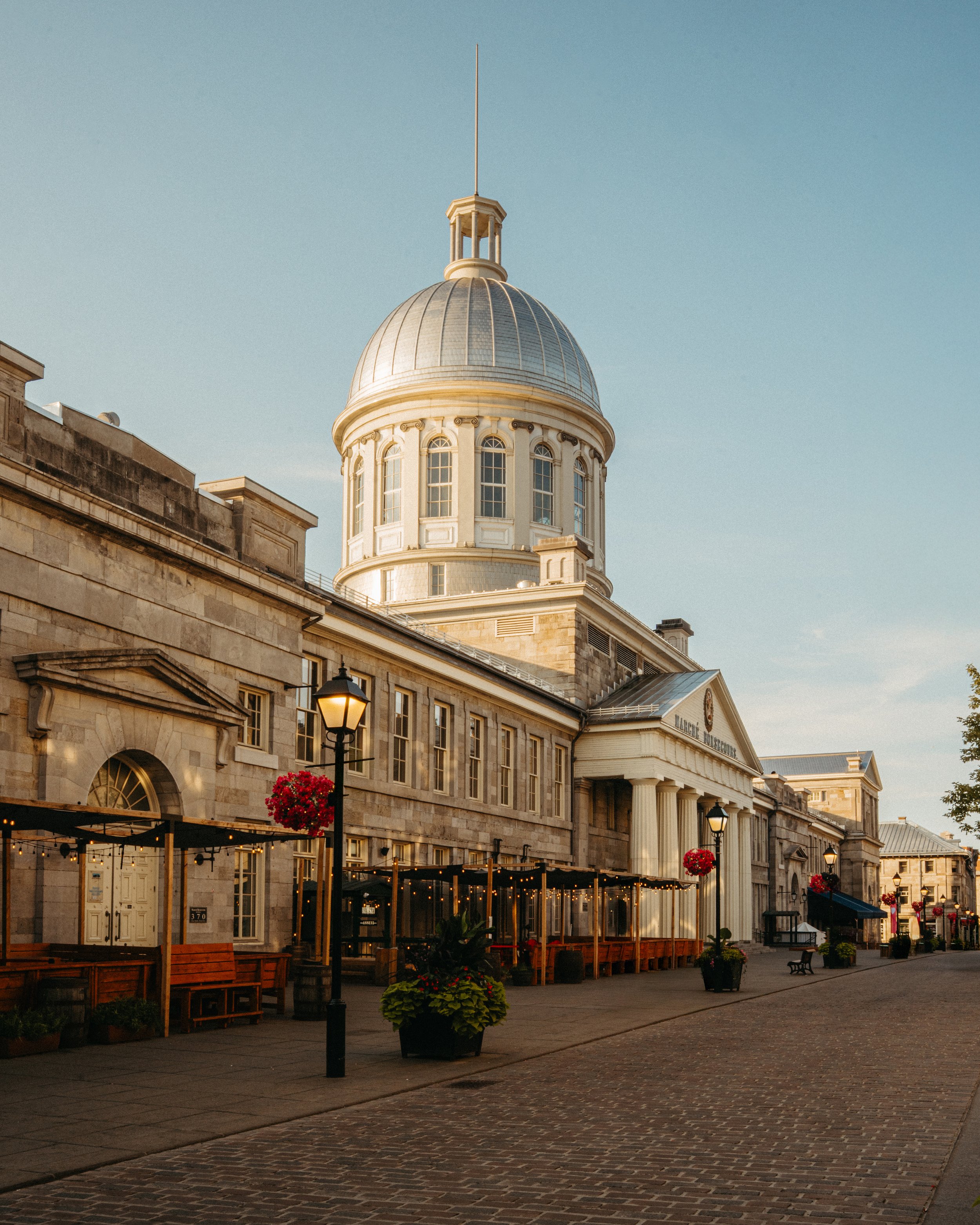 Exterior view of a historic building with a large dome on top, illuminated by soft evening light, with string lights and benches in the foreground.