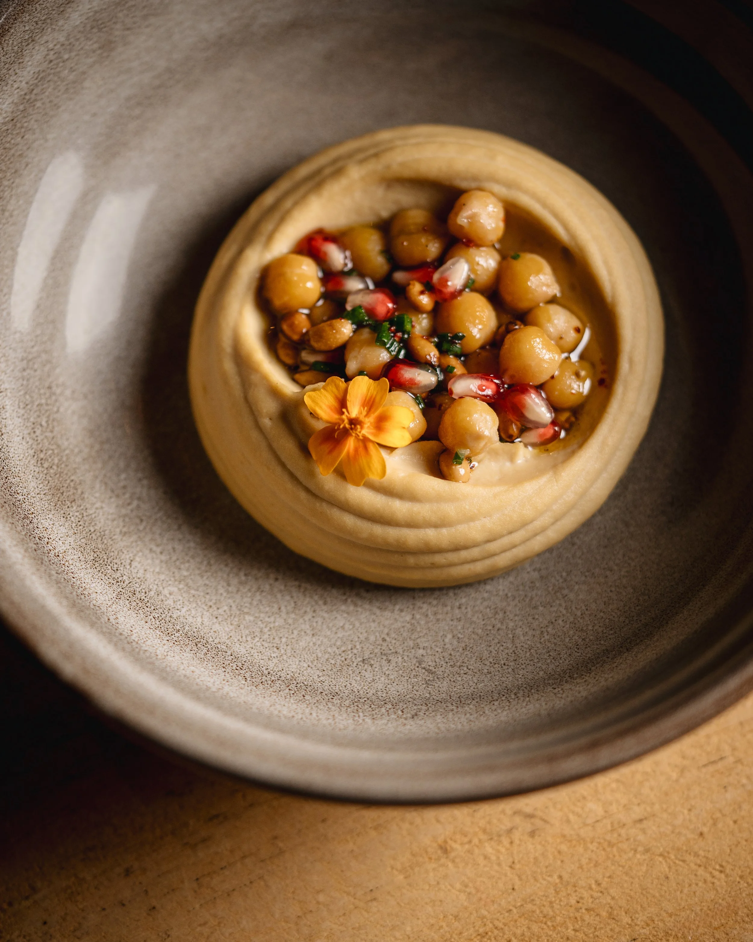 A top view of a round pastry with chickpeas and pomegranate seeds topped with a small yellow flower inside a bowl.