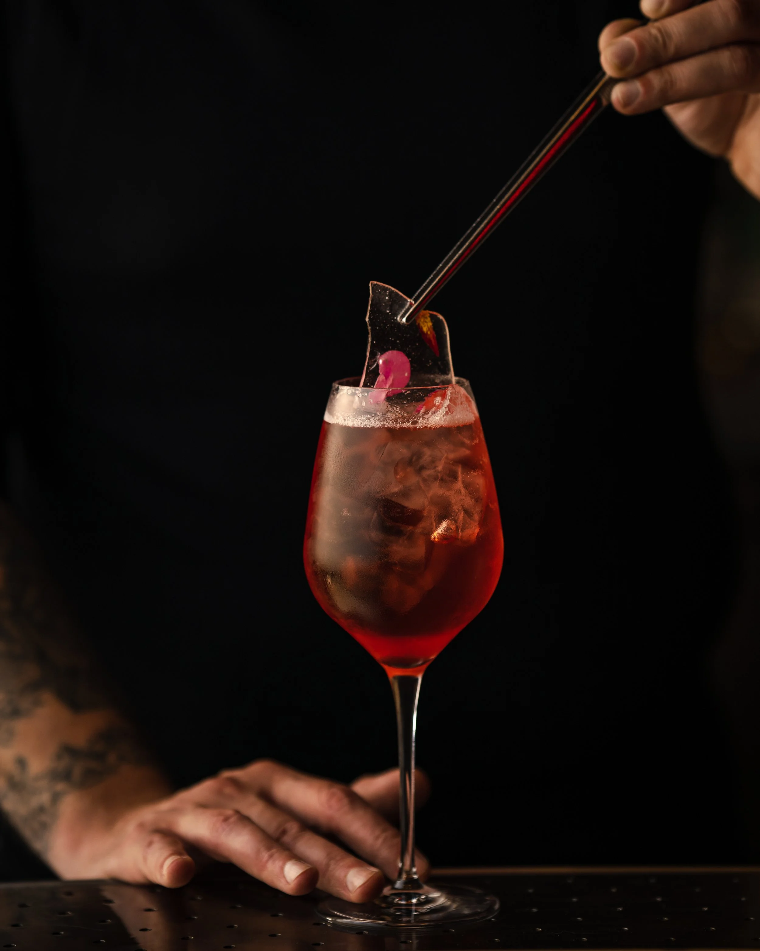 A bartender's hand is seen placing a decorative chocolate garnish onto a cocktail glass filled with a red drink, ice cubes, and garnishes, against a dark background.