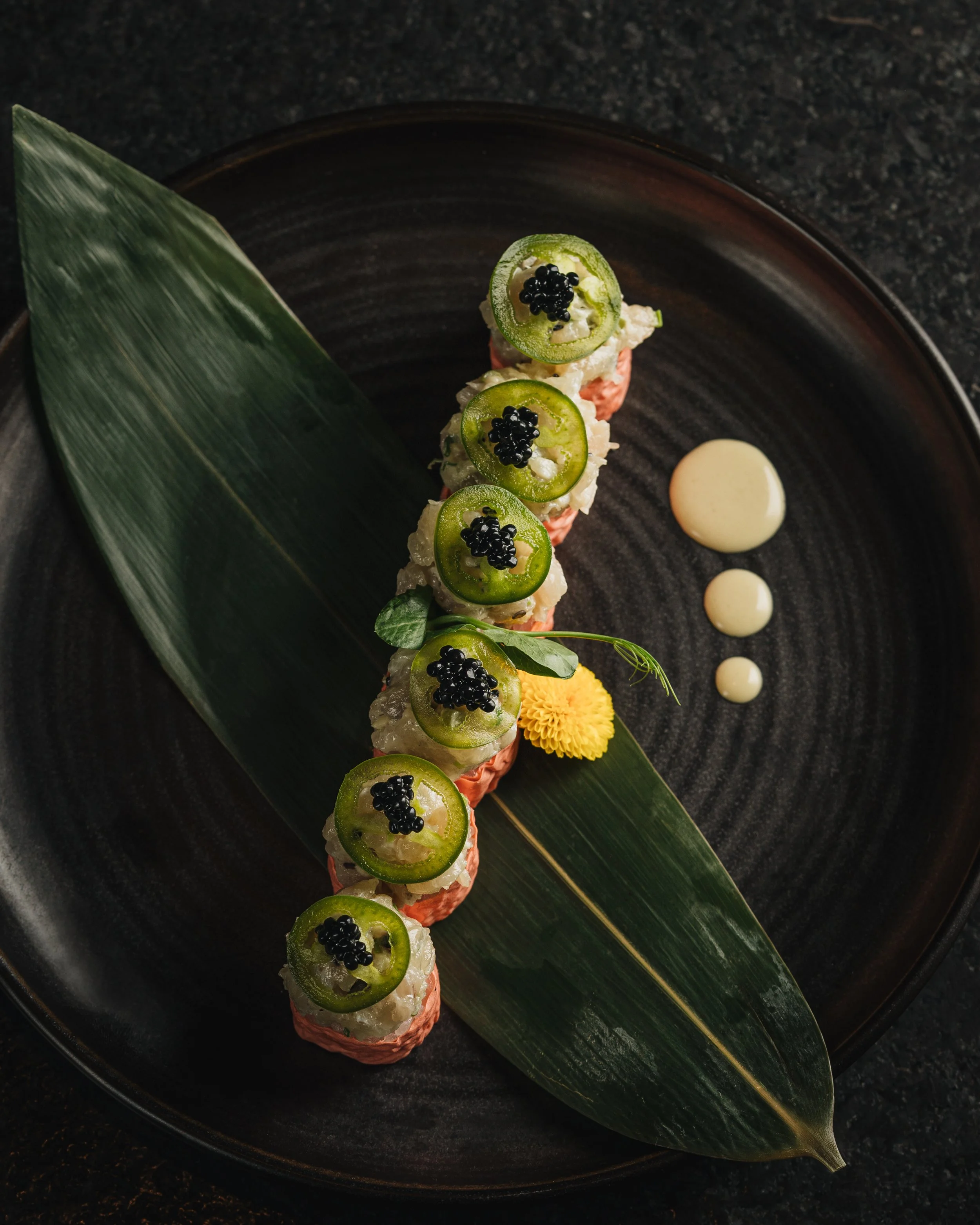 A gourmet sushi roll topped with slices of green jalapeño pepper and black caviar, served with a drizzle of white sauce on a black plate decorated with a large green leaf and a yellow edible flower.