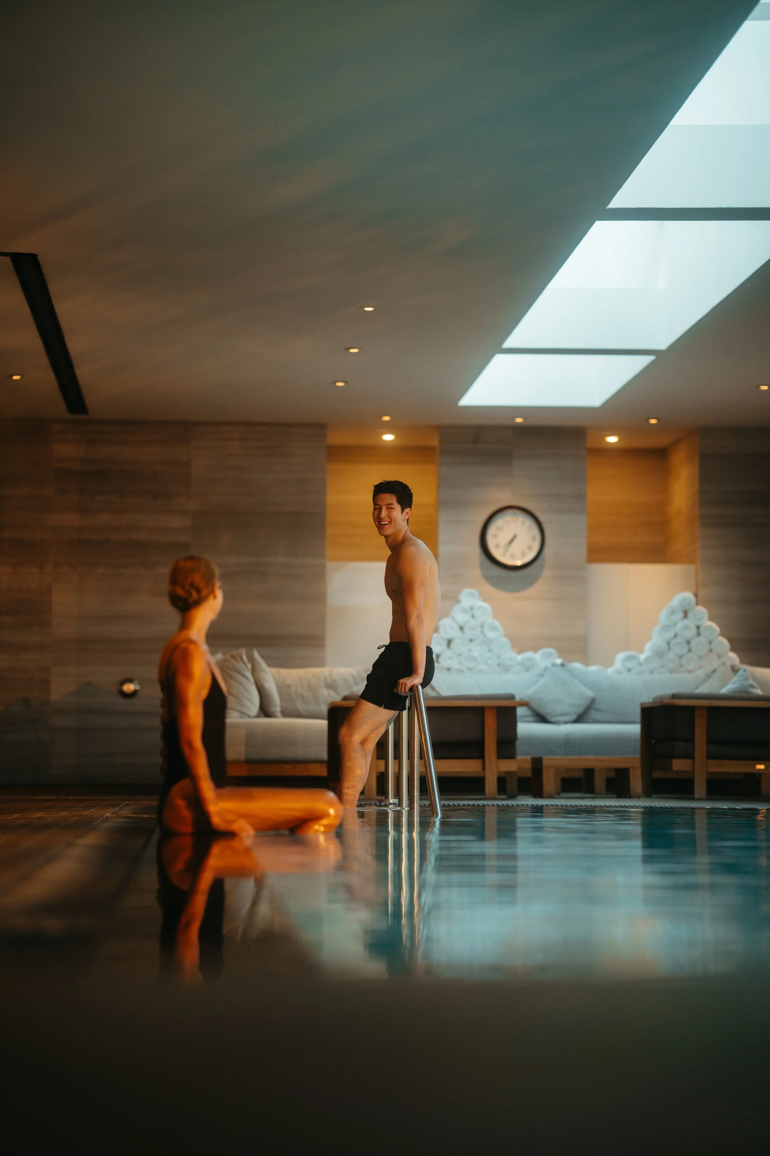 Two people in swimwear by an indoor pool, with one sitting on the floor and the other standing on stairs, smiling and engaging in conversation.