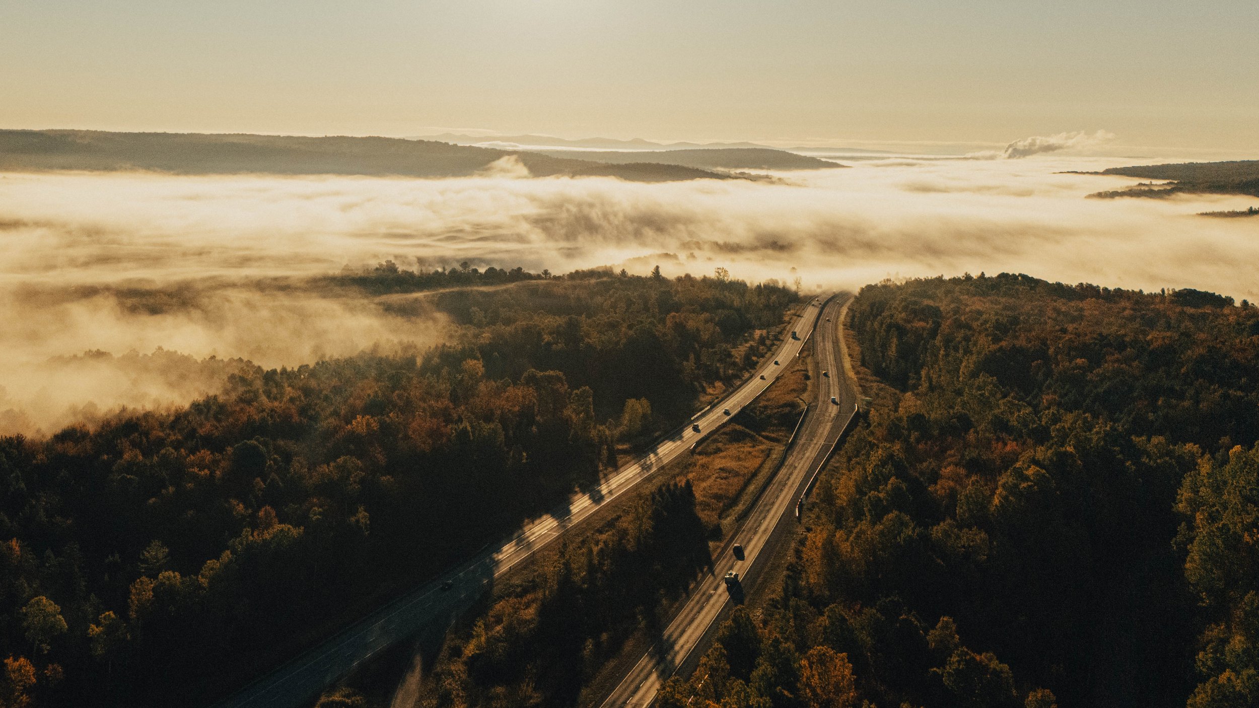 Aerial view of a highway winding through a forested area with hills and mountains in the background, fog or low clouds covering the landscape, during sunrise or sunset.