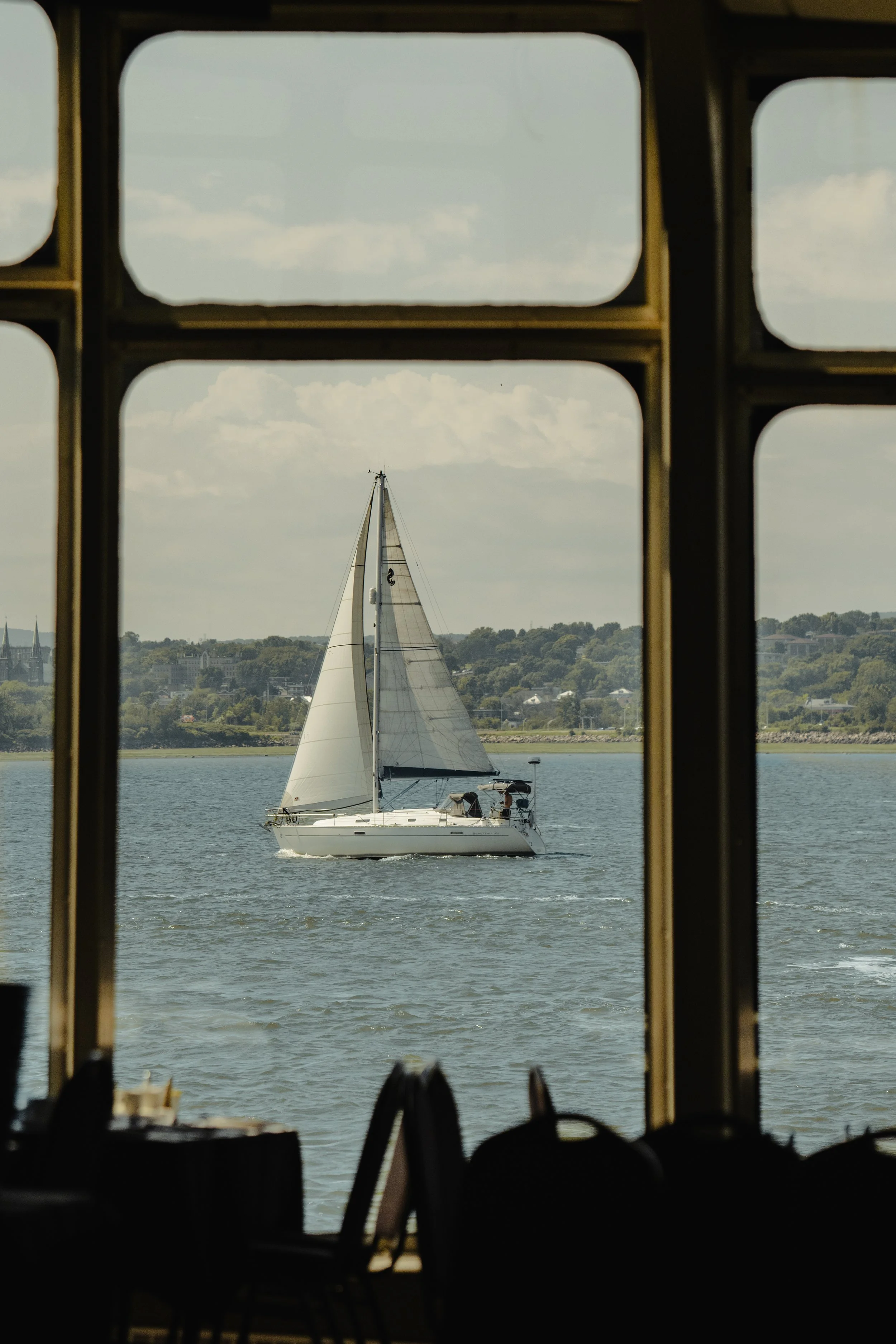 A sailboat on the water viewed through large window panes of a room, with a distant shoreline and hills in the background.