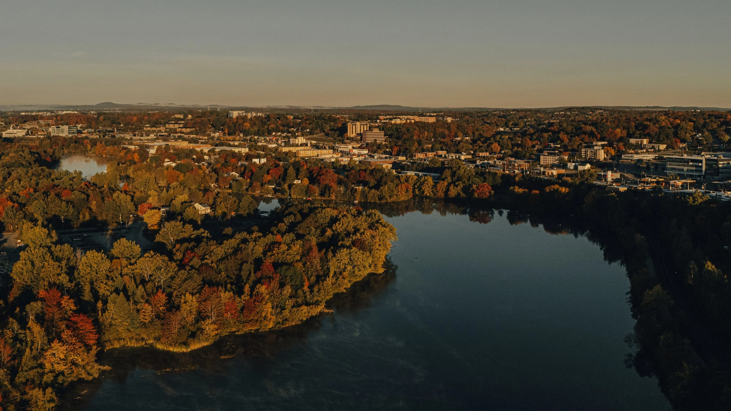 An aerial view of a city with a river, surrounded by trees with autumn foliage, buildings, and industrial areas under a clear sky.