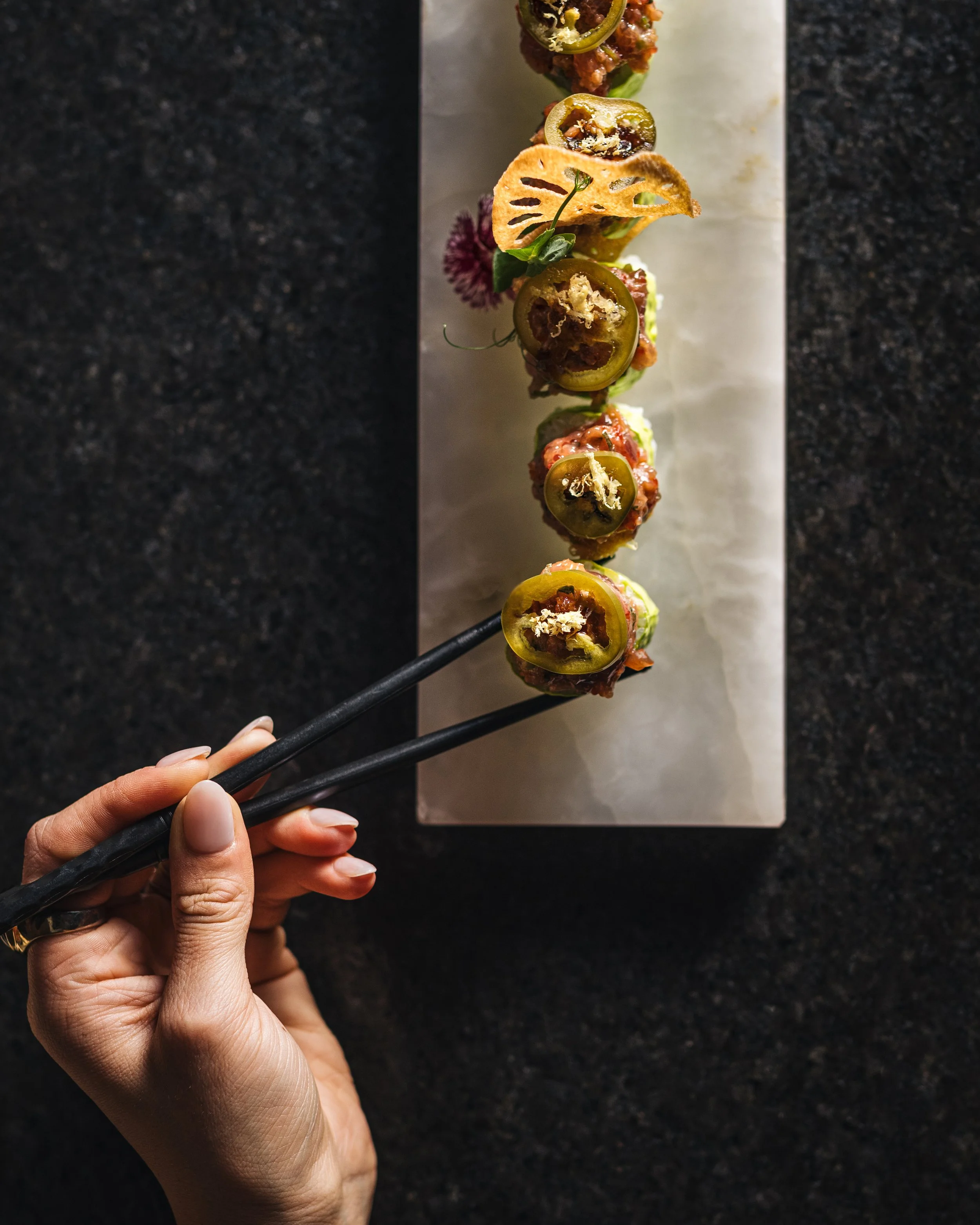 Hand holding black chopsticks over a dish of sushi rolls topped with jalapeño slices and garnished with a purple flower, on a white rectangular plate on a dark countertop.