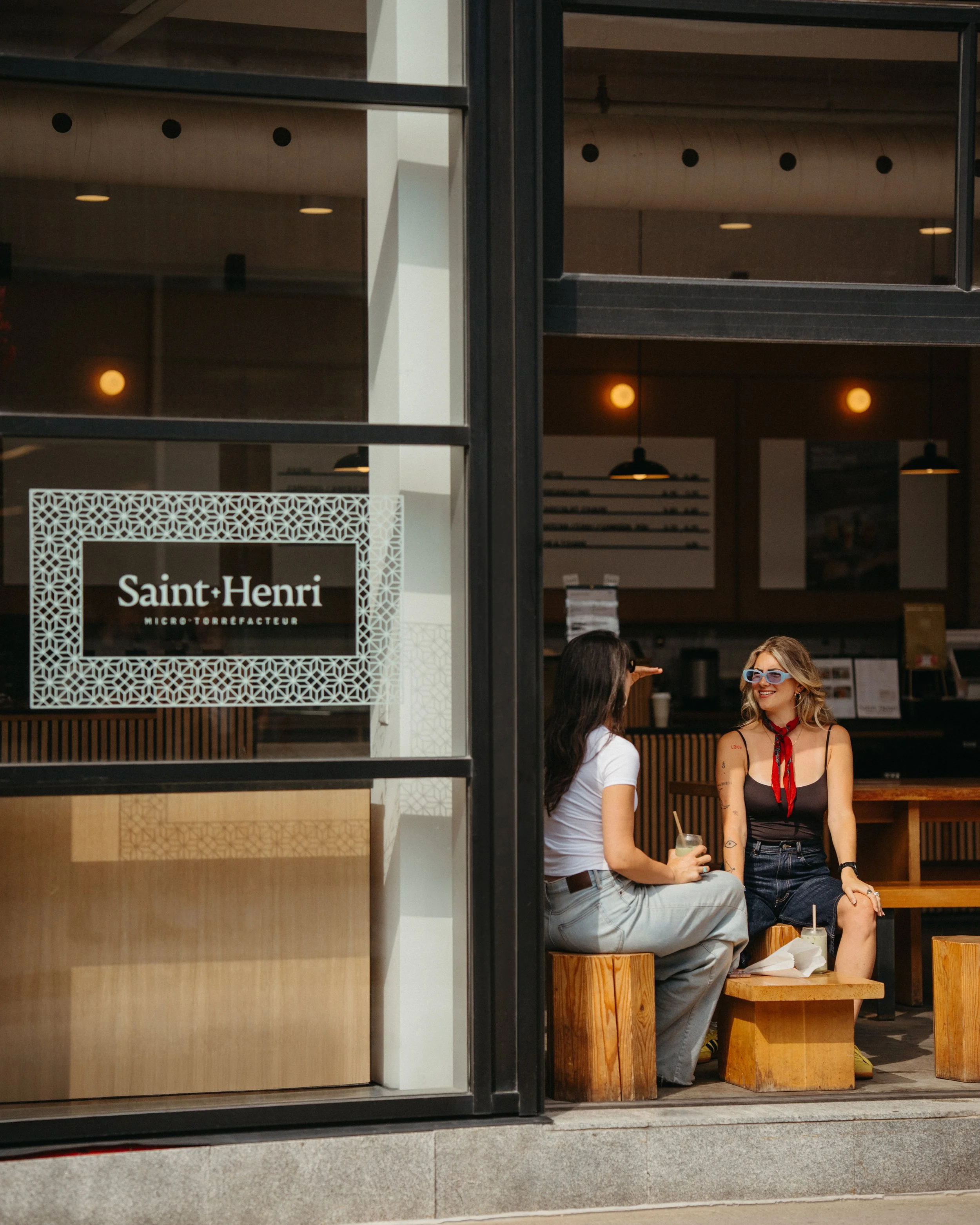 Two women sitting on wooden benches outside a cafe, engaging in conversation. One woman is wearing sunglasses, a black top, and has tattoos on her arms, holding a drink. The other is wearing a white top, and her face is not visible. The cafe sign rea