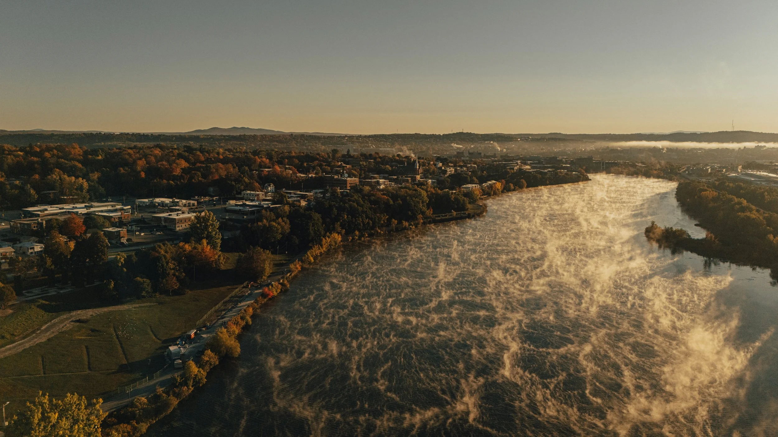 Aerial view of a river flowing through a city surrounded by trees with autumn foliage, buildings, and hills in the background during sunset or sunrise.