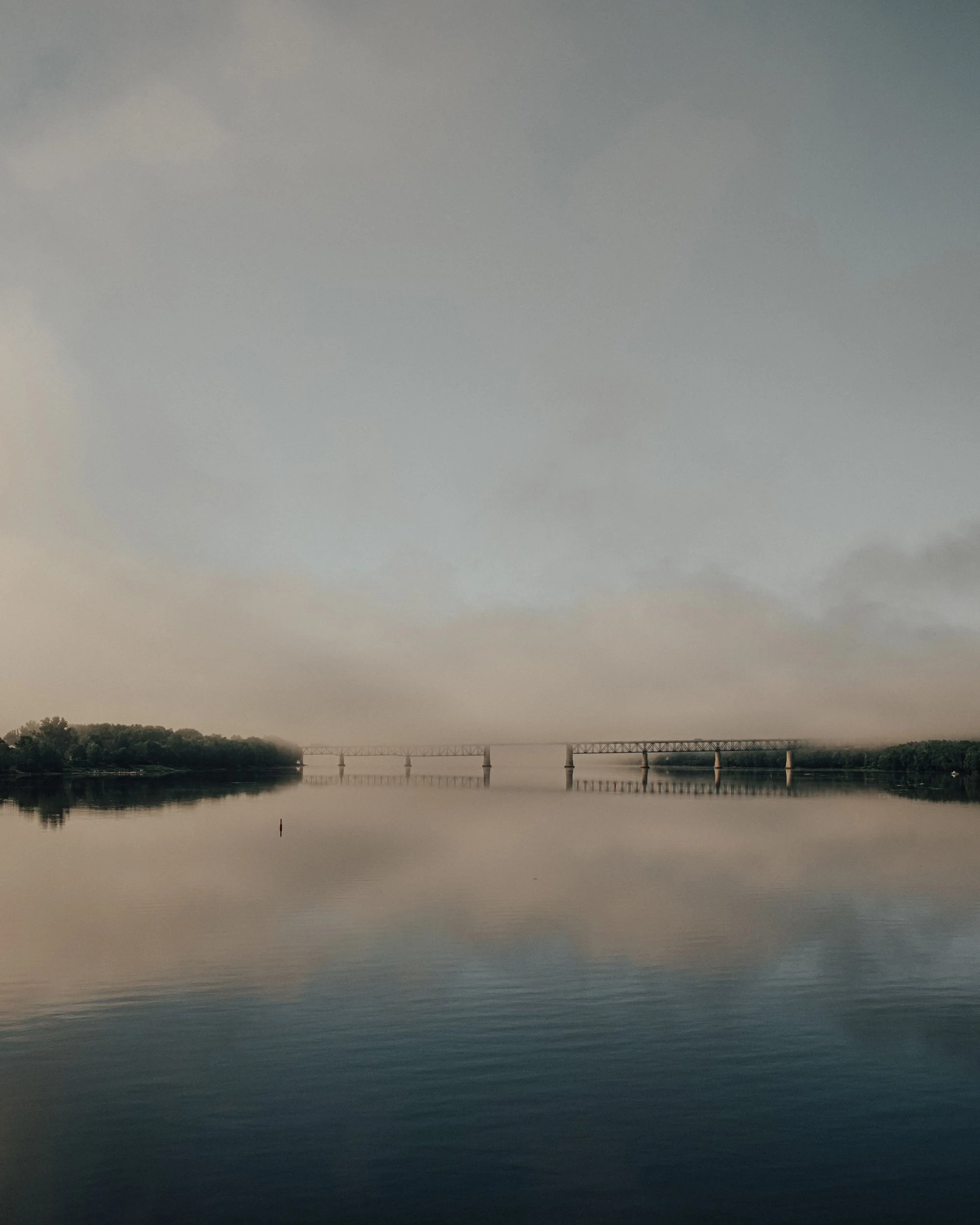 A calm river under a cloudy sky, with trees on the shoreline left and a bridge in the distance.