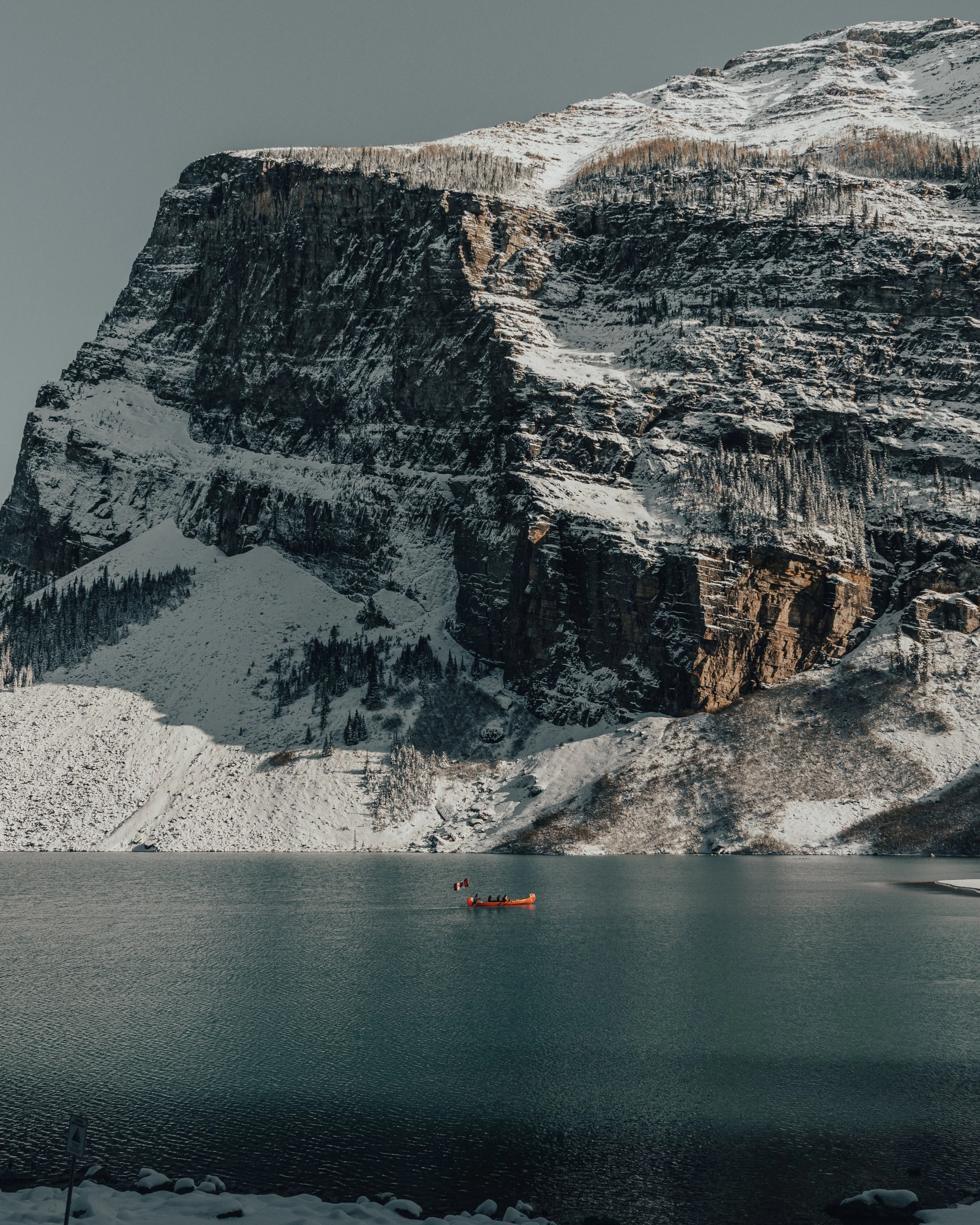 Snow-covered mountain with steep cliffs, trees, and a snowfield at the base, with a peaceful lake in the foreground and a red boat with people on the water.
