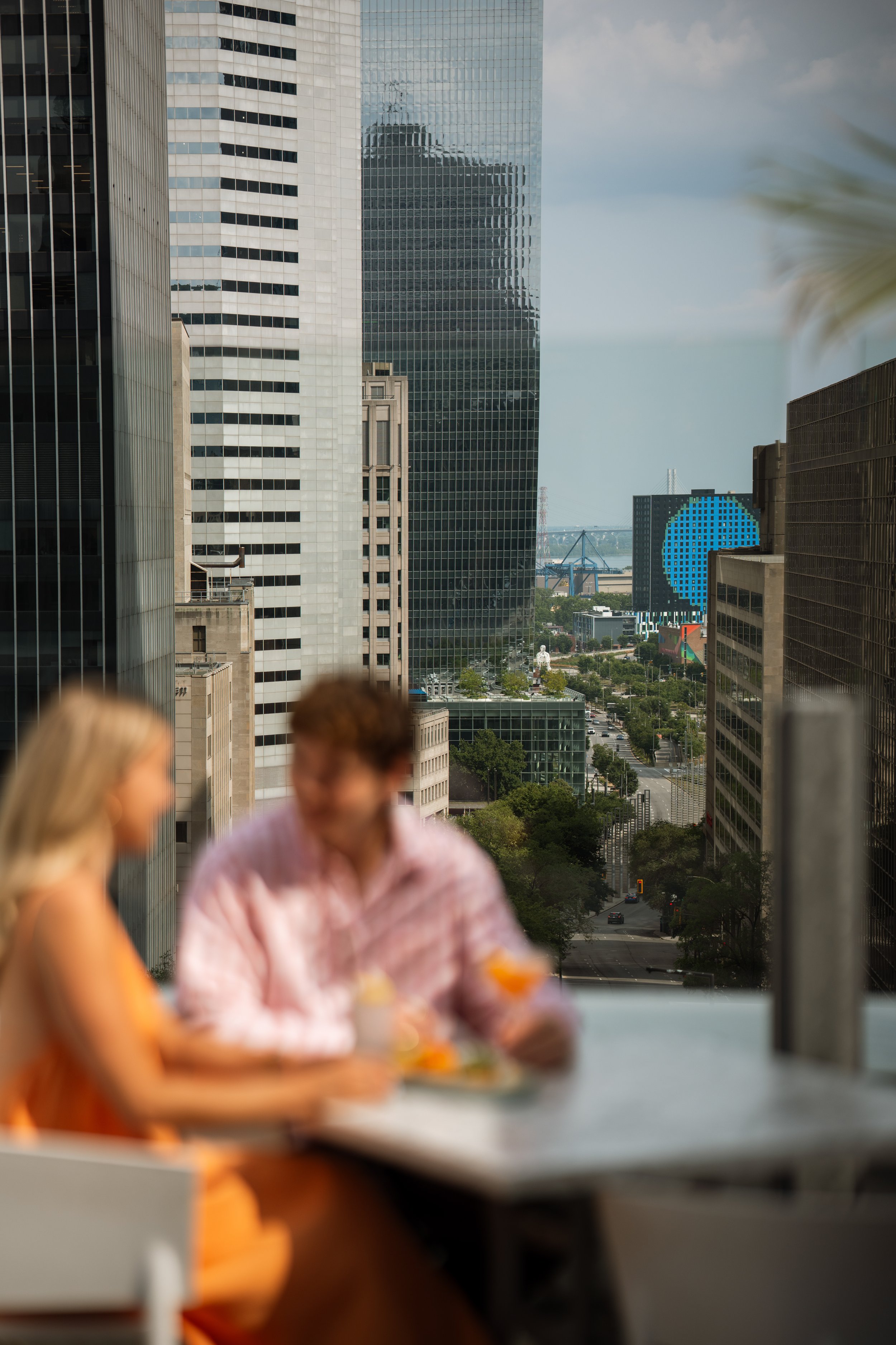 A cityscape with tall glass skyscrapers and a view of a bridge in the distance, seen from a rooftop or balcony with people in the foreground.