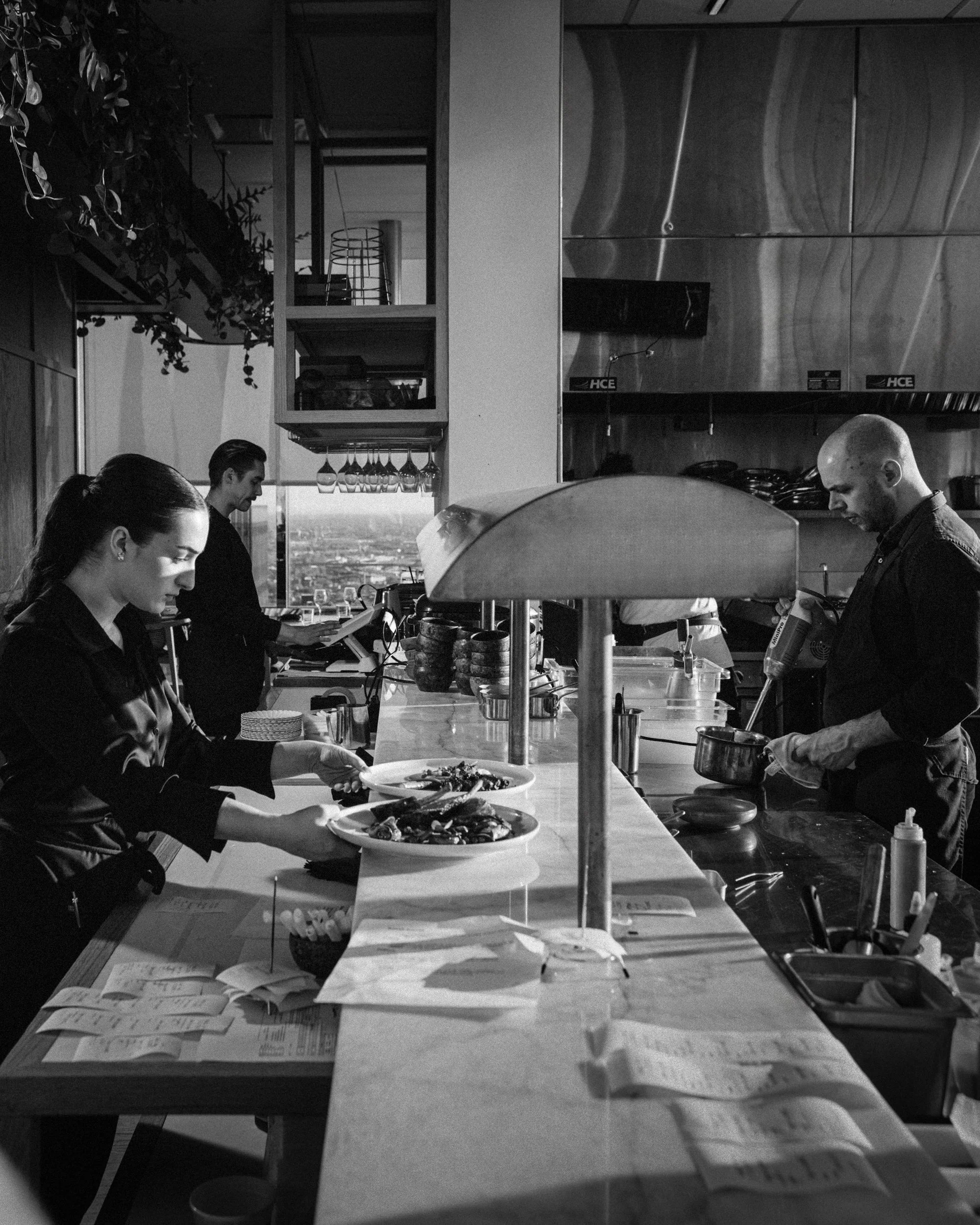 People working in a restaurant kitchen preparing food, with menus and utensils on the counter, and shelves with glasses and kitchen tools in the background.