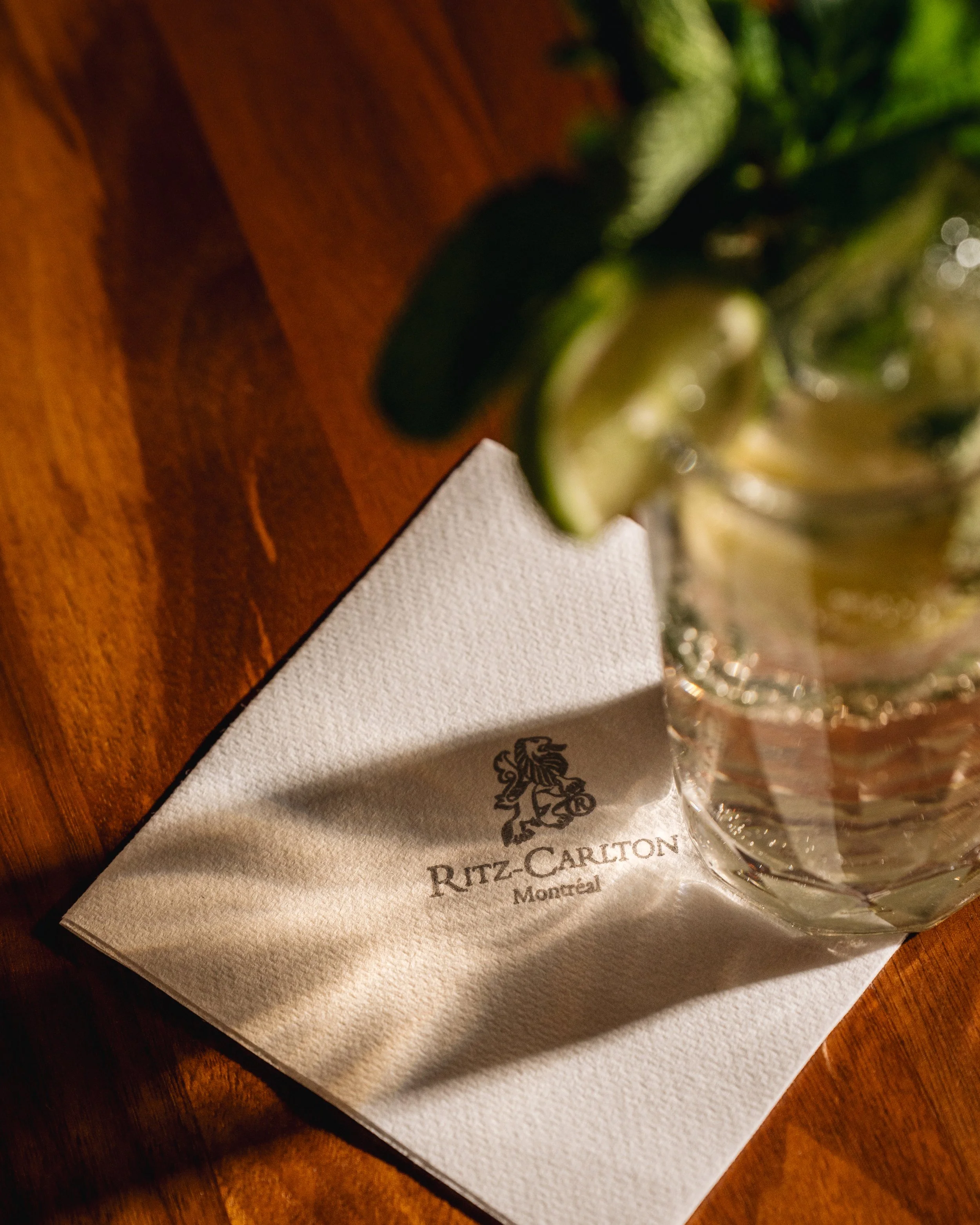 A close-up view of a Ritz-Carlton napkin with the hotel logo and location in Montreal, placed on a wooden table. A water glass and a blurred plate of food are also visible in the background.