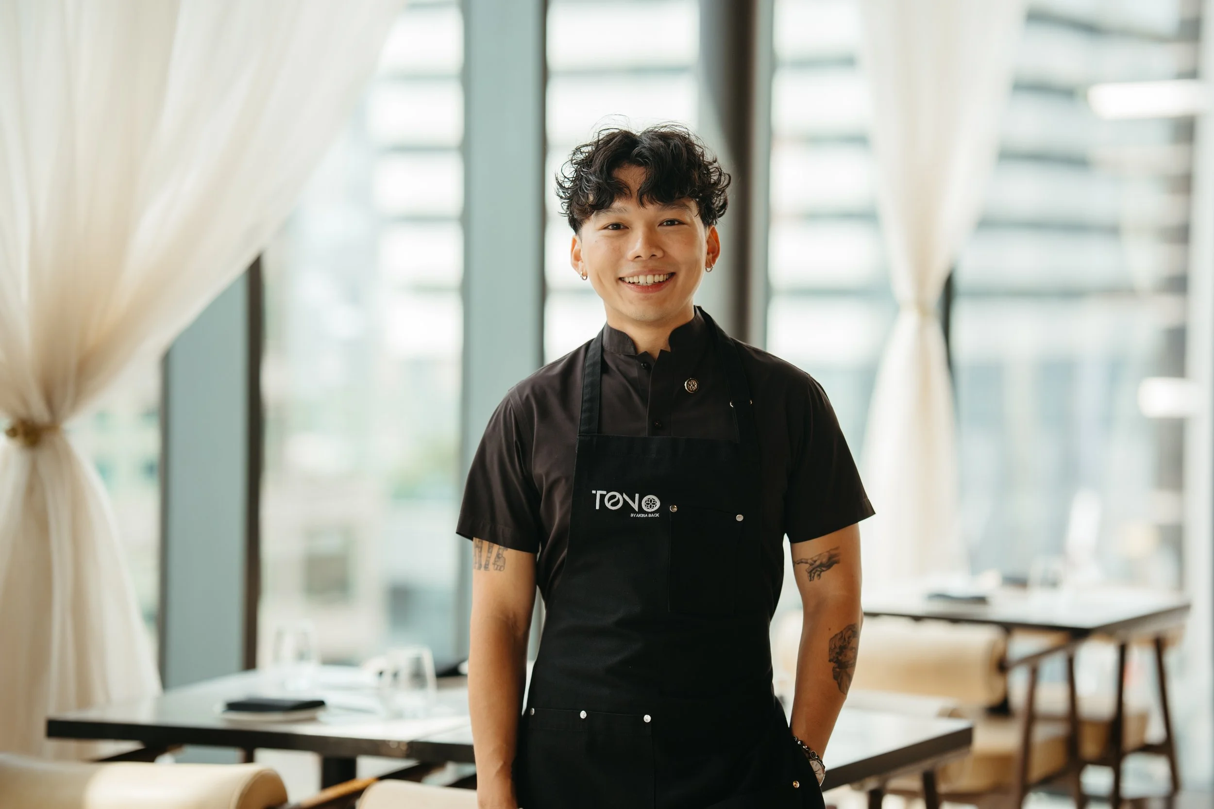 A smiling man with dark curly hair, earrings, and tattoos on his arms, wearing a black shirt and apron, standing inside a bright, modern restaurant with large windows and curtains in the background.
